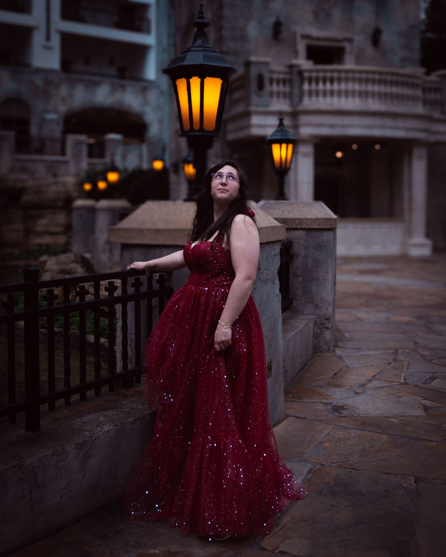 Woman in sparkling red ball gown posing by stone railing with glowing lanterns at dusk.