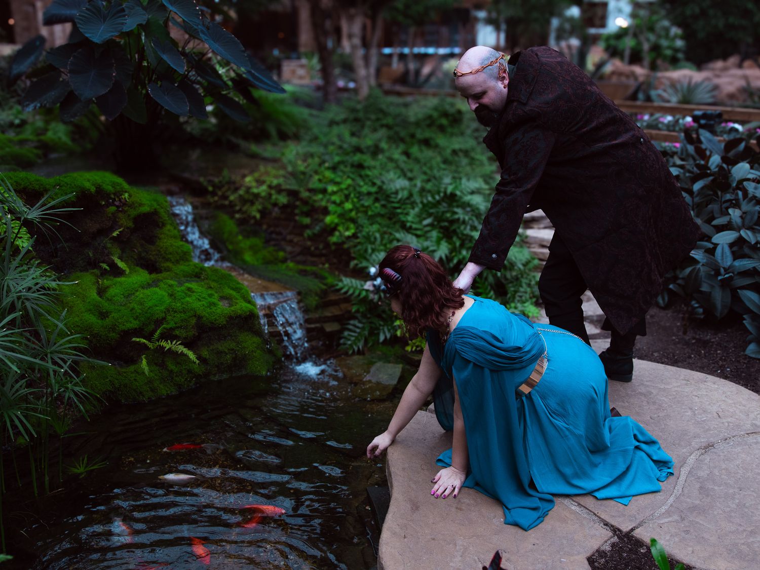 Two costumed performers pose by a koi pond in a lush garden setting, one kneeling in a teal gown.