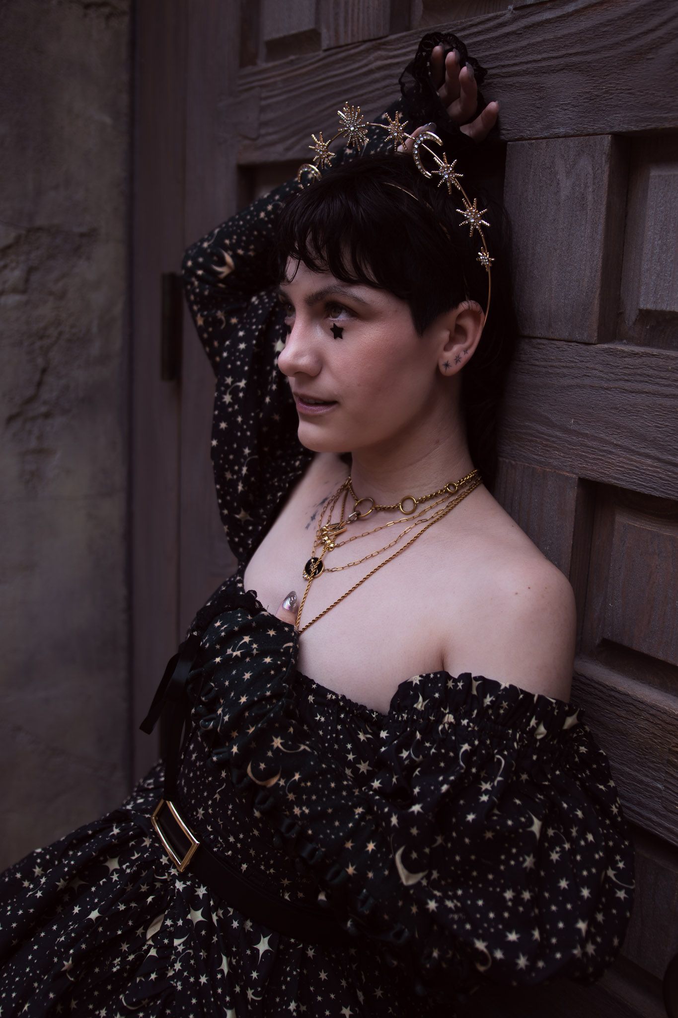 Woman in black star-print off-shoulder dress with gold celestial headpiece and layered necklaces leaning against wooden door.