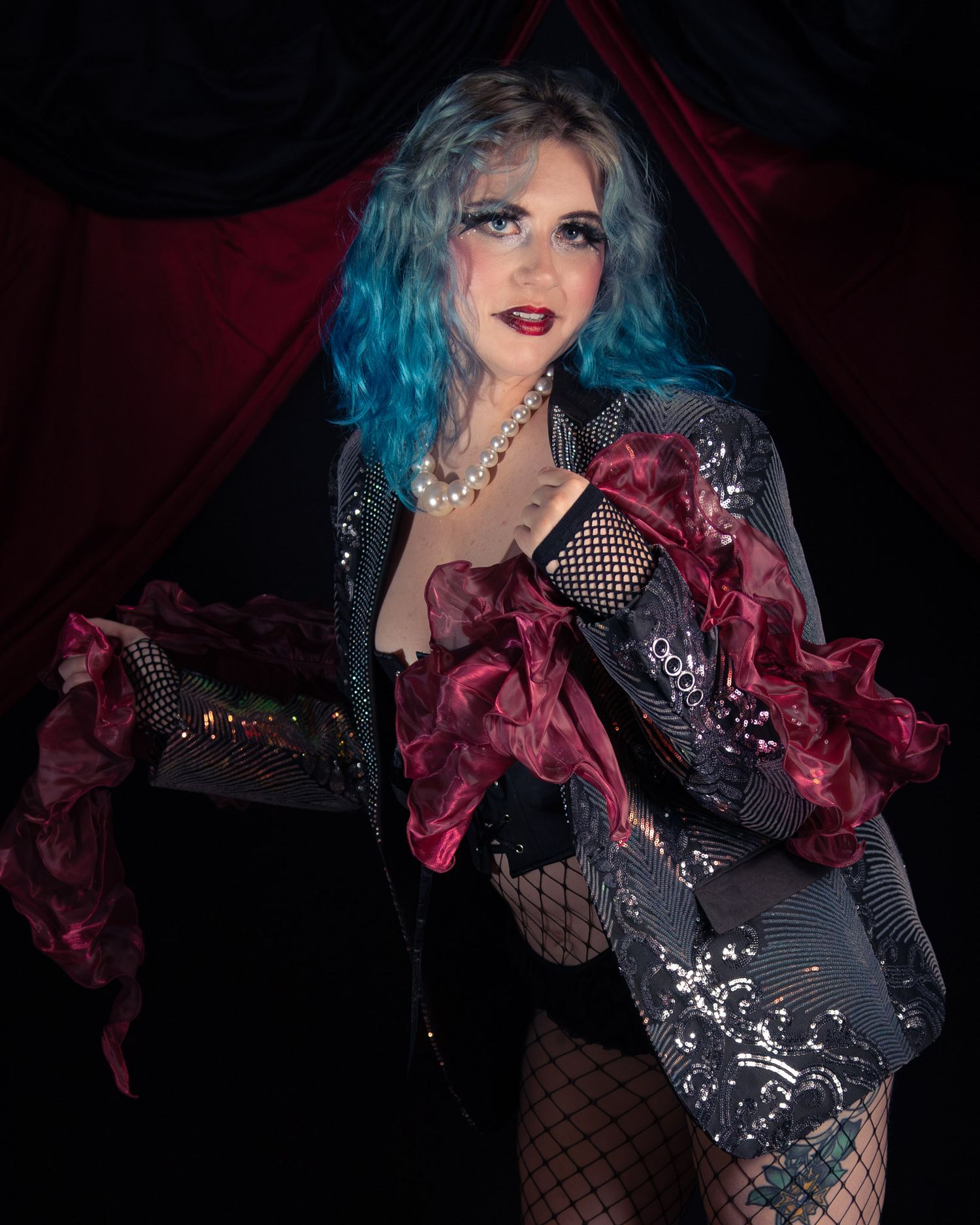 A performer in blue hair, fishnet stockings, sequined jacket, and burgundy ruffled accessories poses dramatically on a dark stage.