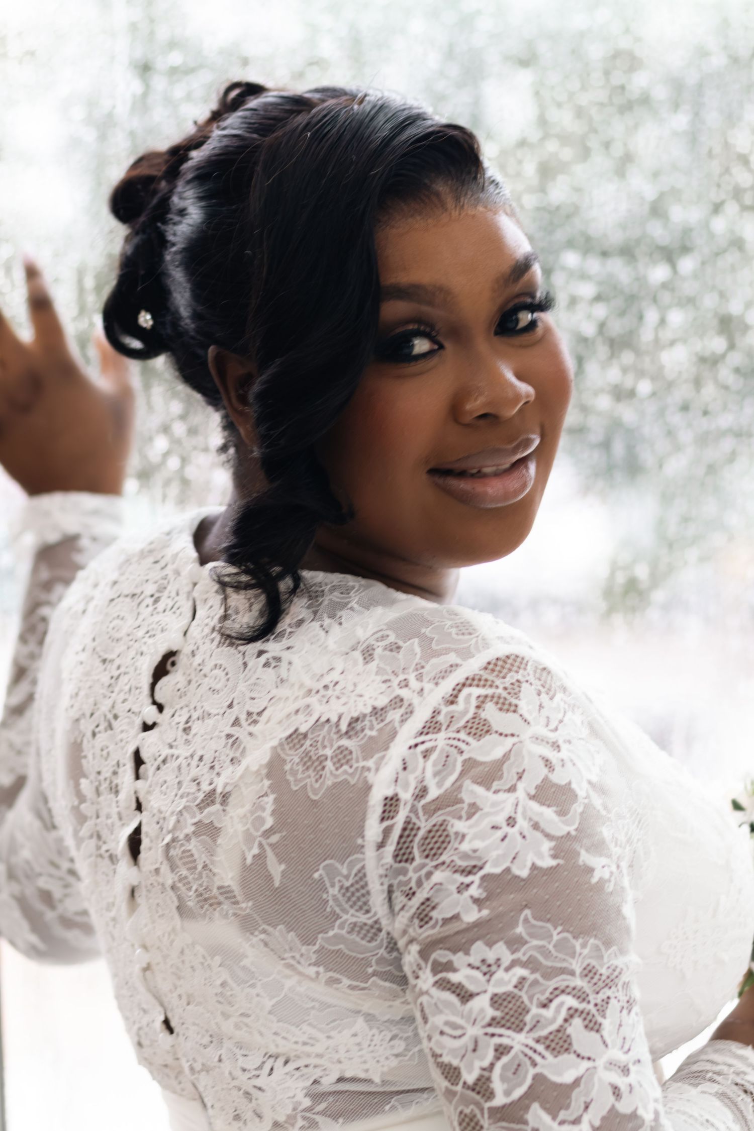 A smiling bride in a white lace long-sleeve gown poses elegantly, looking over her shoulder near a rain-covered window.