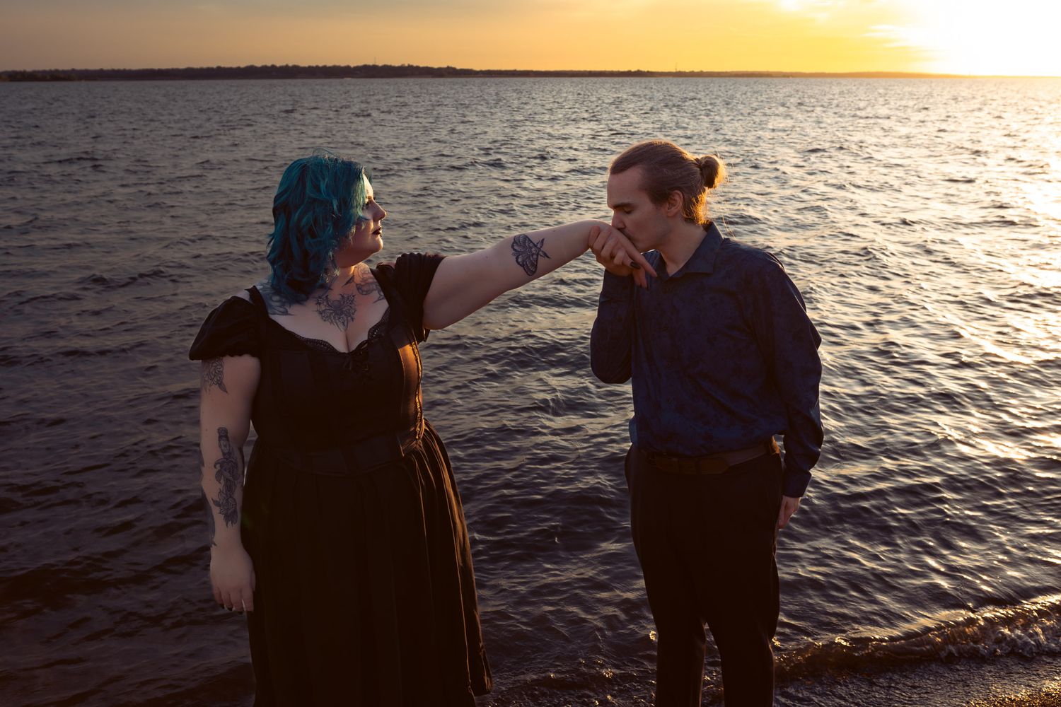 A man kisses a tattooed woman with blue hair's hand by a sunset lake shore.