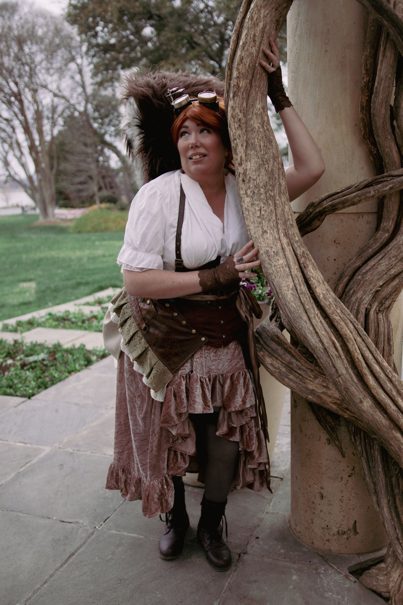 Woman in steampunk costume with corset, ruffled skirt, goggles, and squirrel tail poses by twisted tree outdoors.