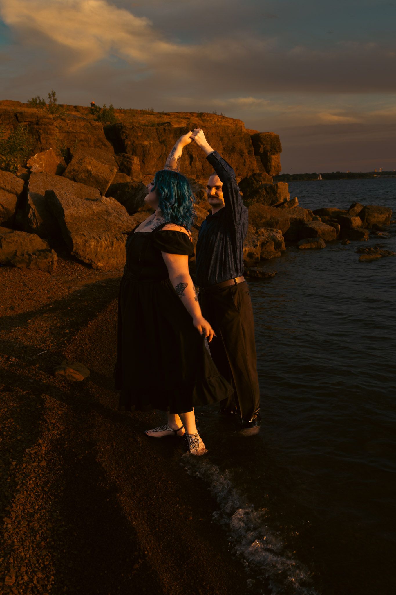Two people dancing on rocky shoreline at sunset, one with blue hair in black dress, waves lapping at their feet.