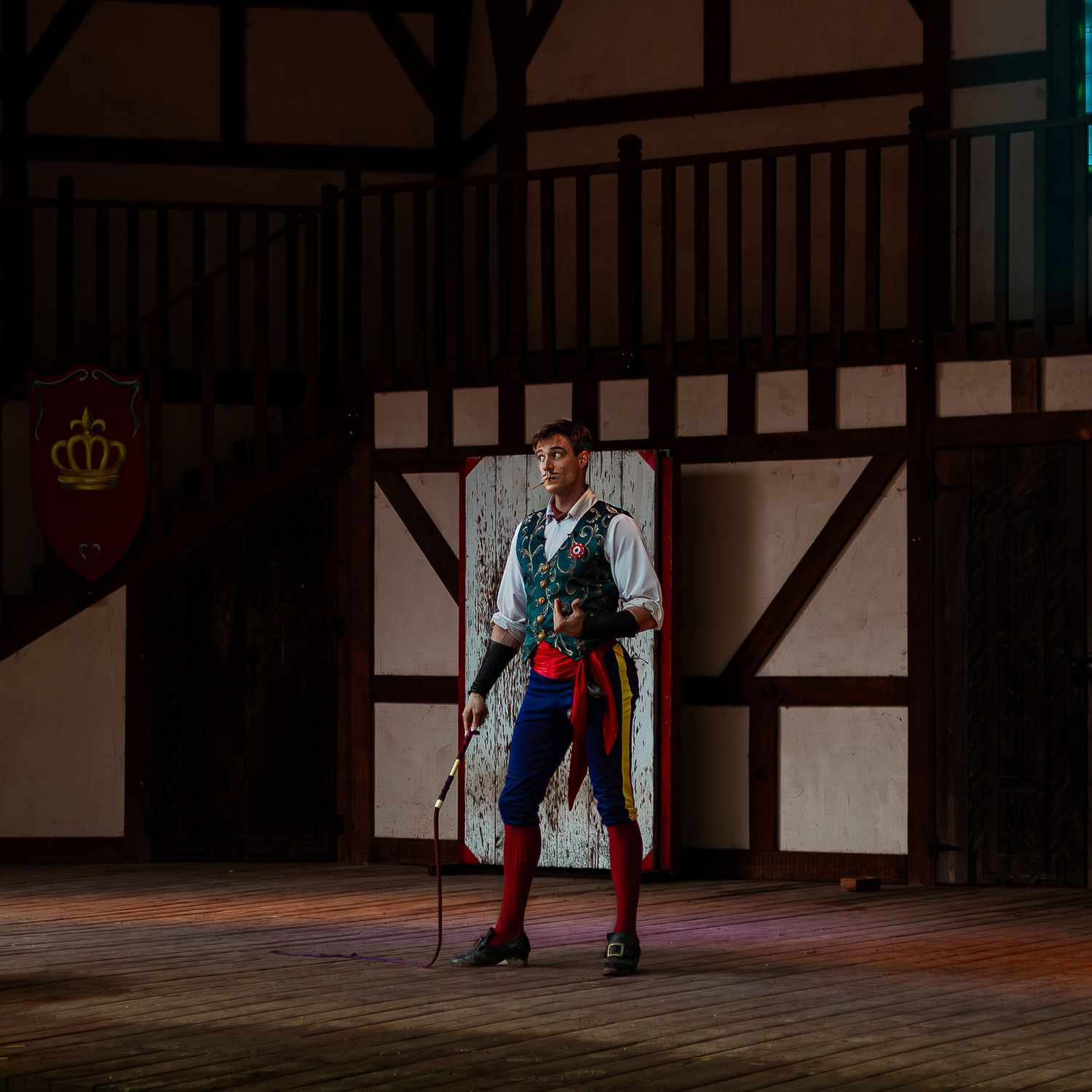 A costumed performer in medieval attire stands on a wooden stage inside a Tudor-style building with a royal crest banner.