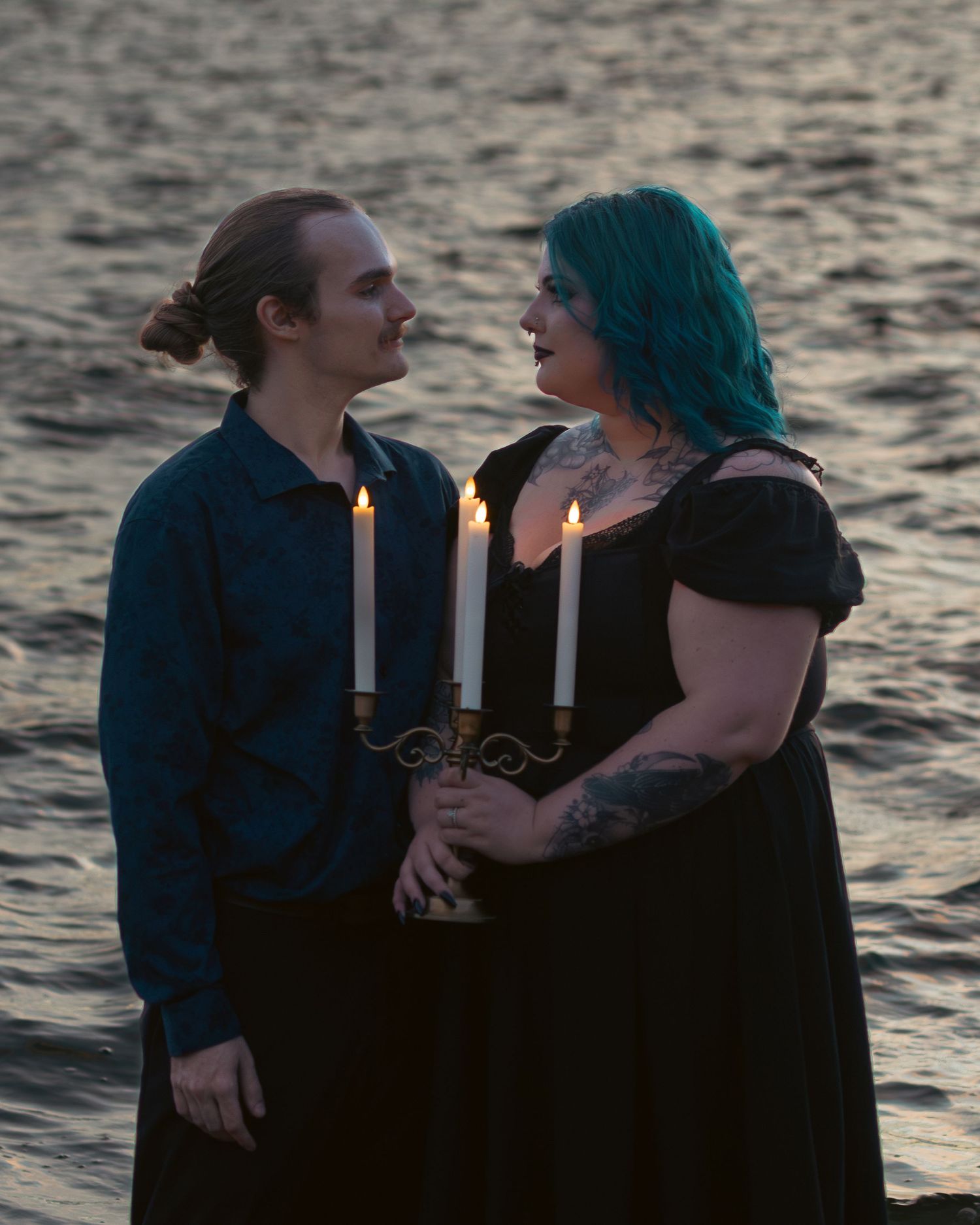 Couple holding candelabra with lit candles near water at dusk, woman with teal hair in black dress, man in dark shirt.