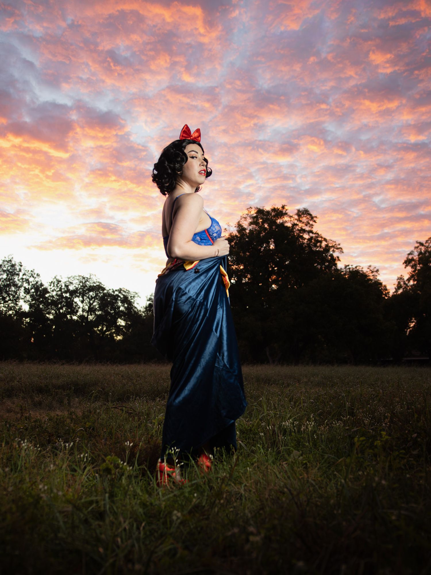 Woman in a Snow White costume with a red bow and blue satin skirt stands in a grassy field beneath a vivid pink and orange sunset sky.