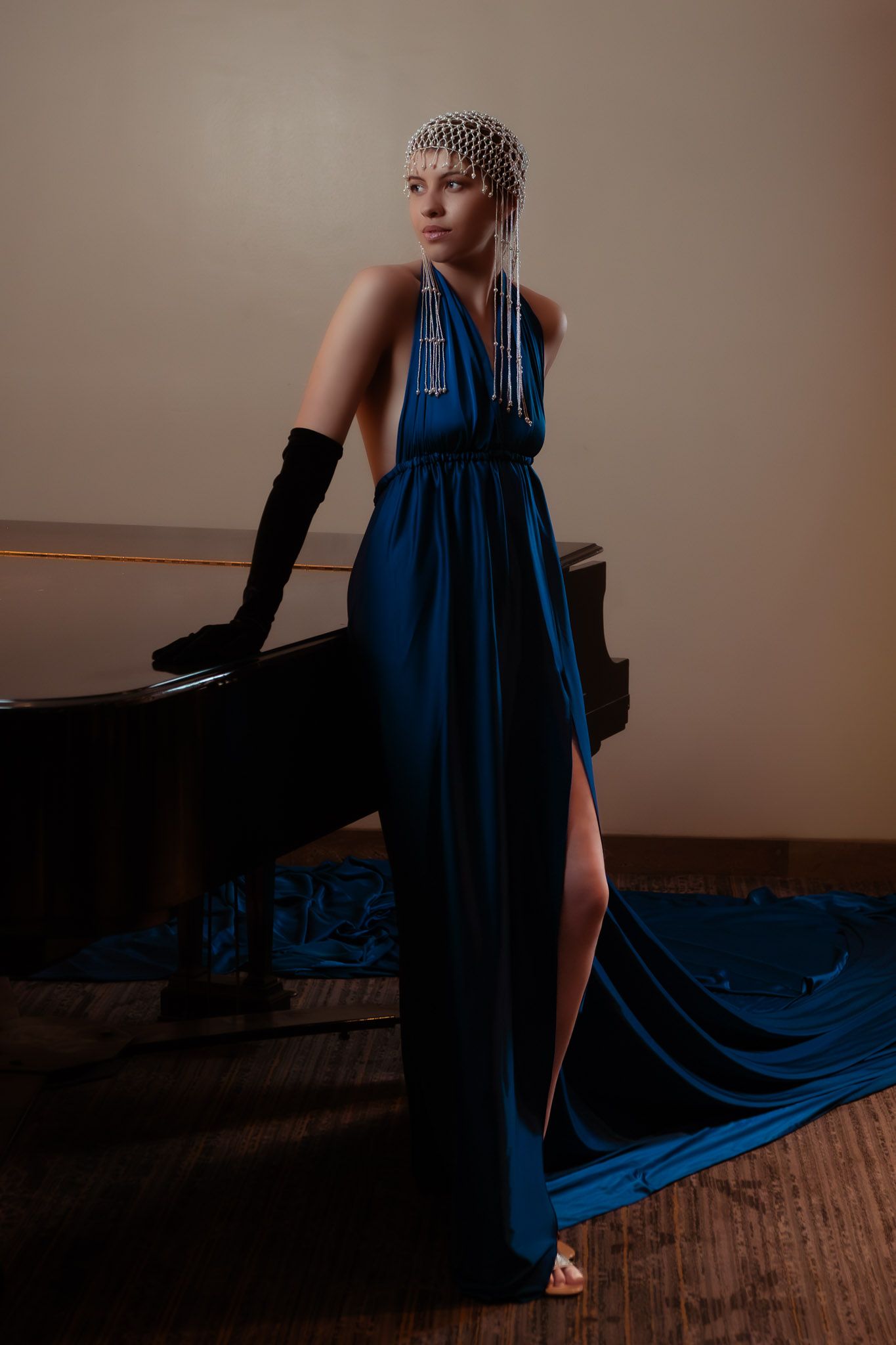 Woman in blue halter gown with pearl headpiece poses elegantly beside a grand piano wearing black gloves.