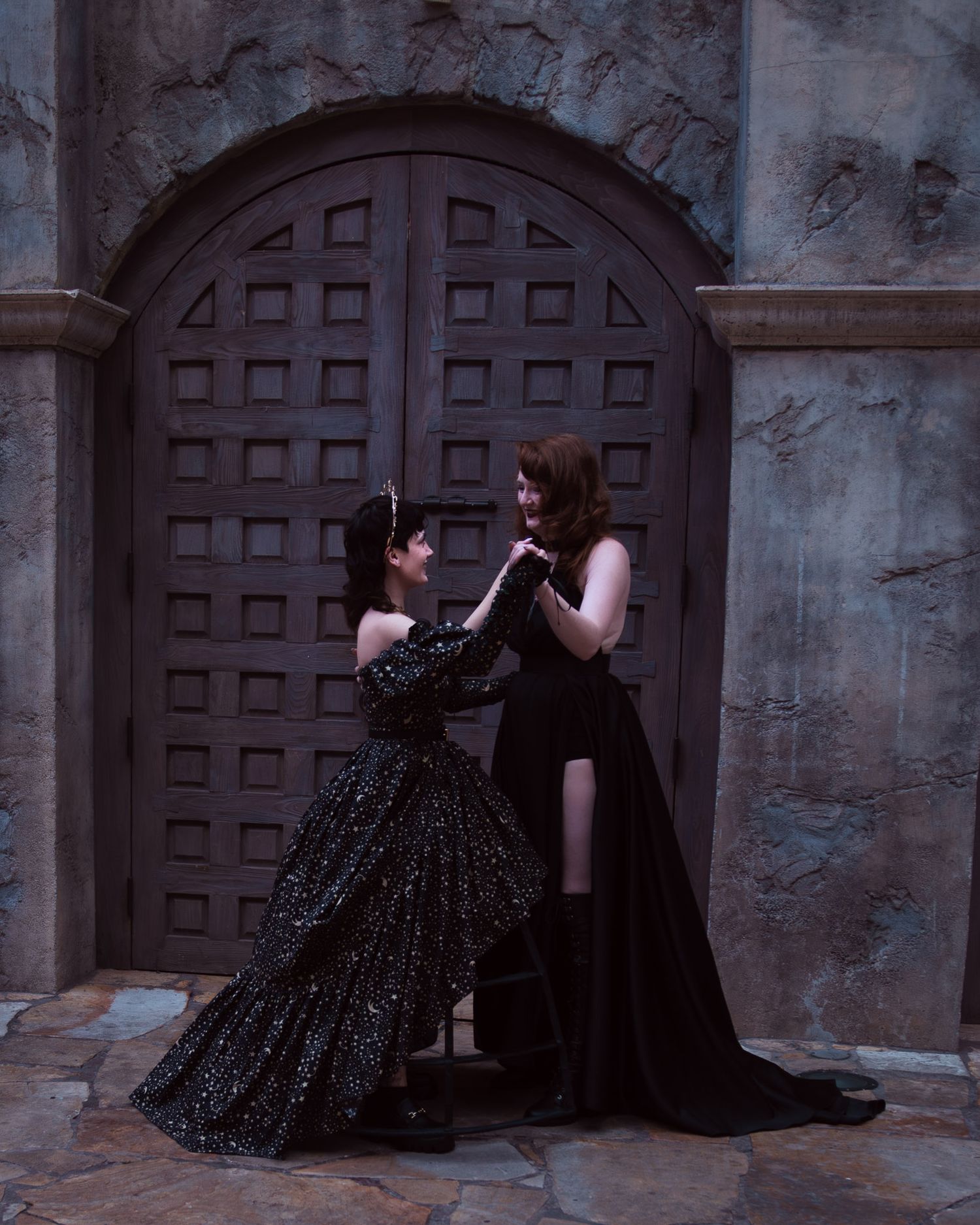 A moody fantasy photo of two women dancing before a large wooden gate. One wears a celestial star-print gown; both smile as they hold hands.
