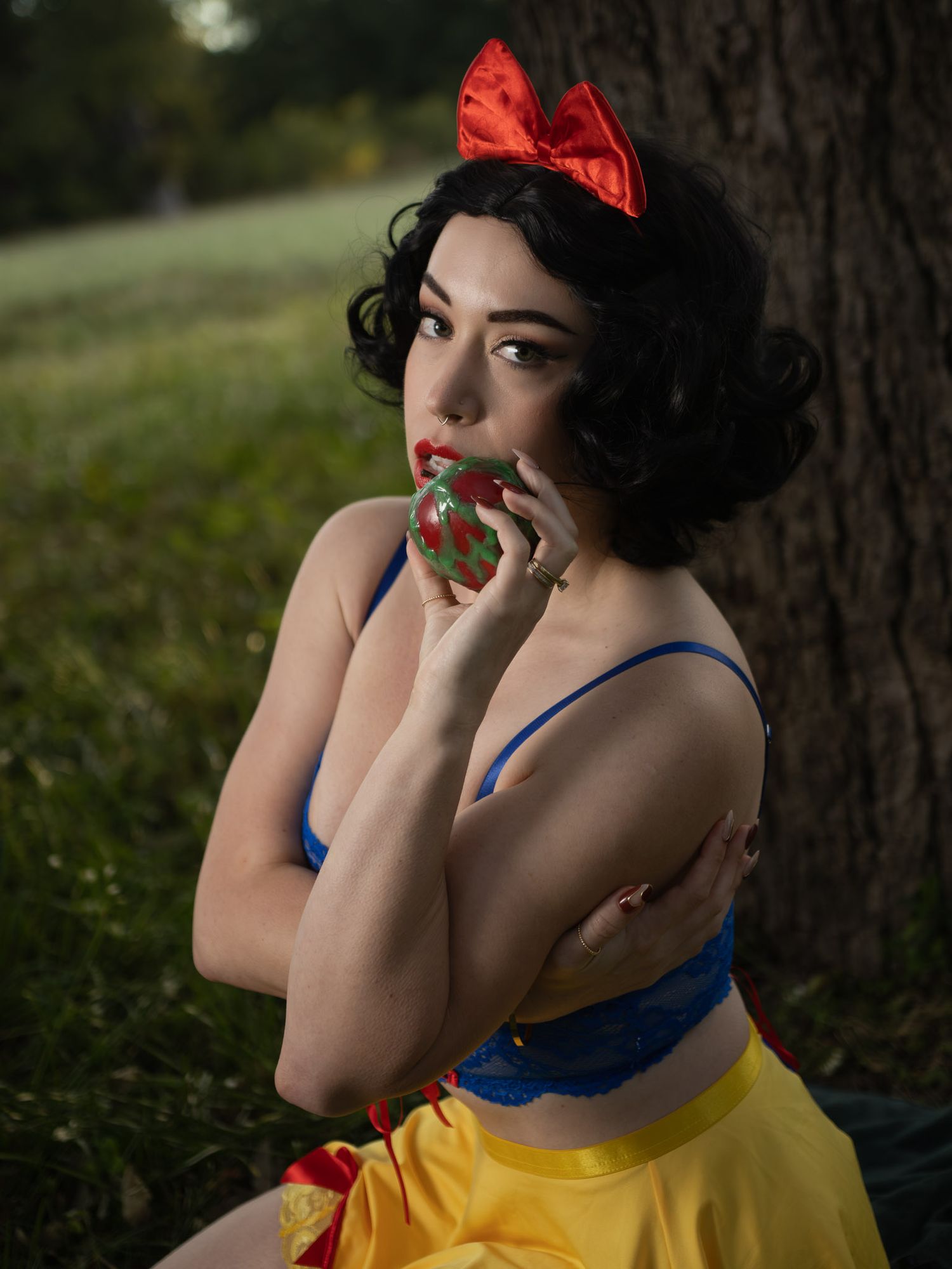 Woman in a Snow White-inspired cosplay with a red bow, blue top and yellow skirt holds a red apple to her lips beside a tree.