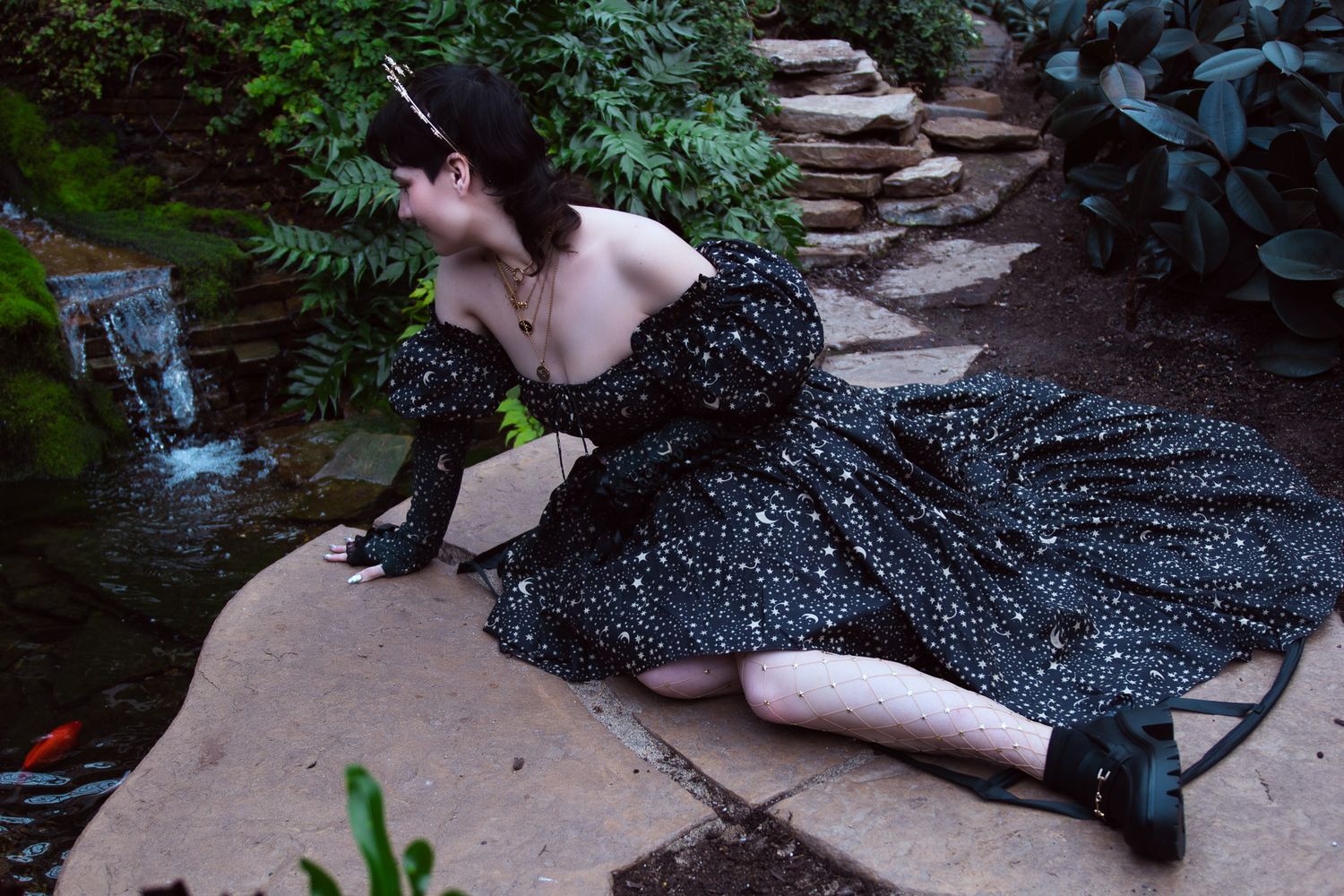 Woman in black starry off-shoulder gown poses gracefully on stone path beside koi pond in lush garden.