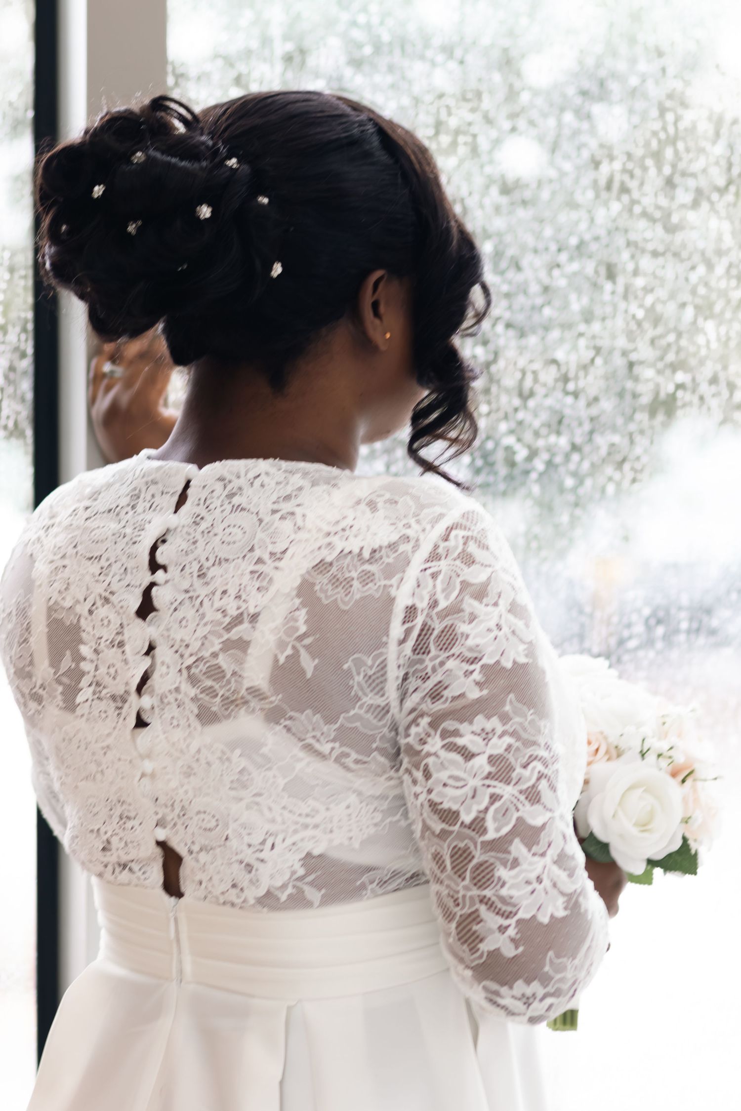 Luminous back view of a bride's wedding gown highlighting intricate lace button details against a bright window.