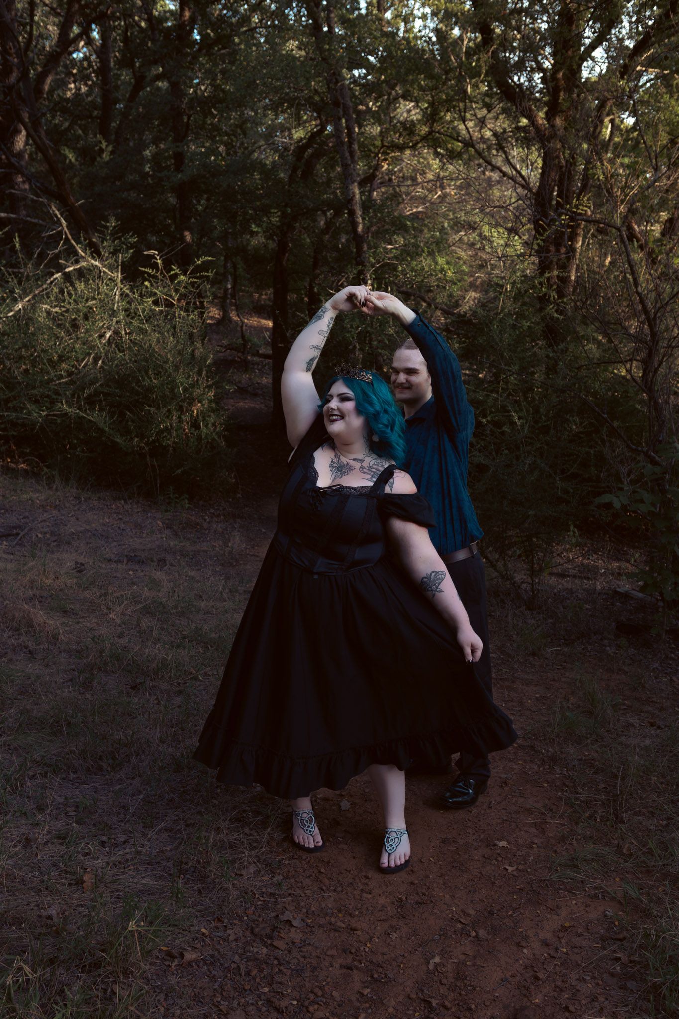 Couple dancing outdoors on a forest path, woman in black dress with teal hair, partner spinning her under raised arms.