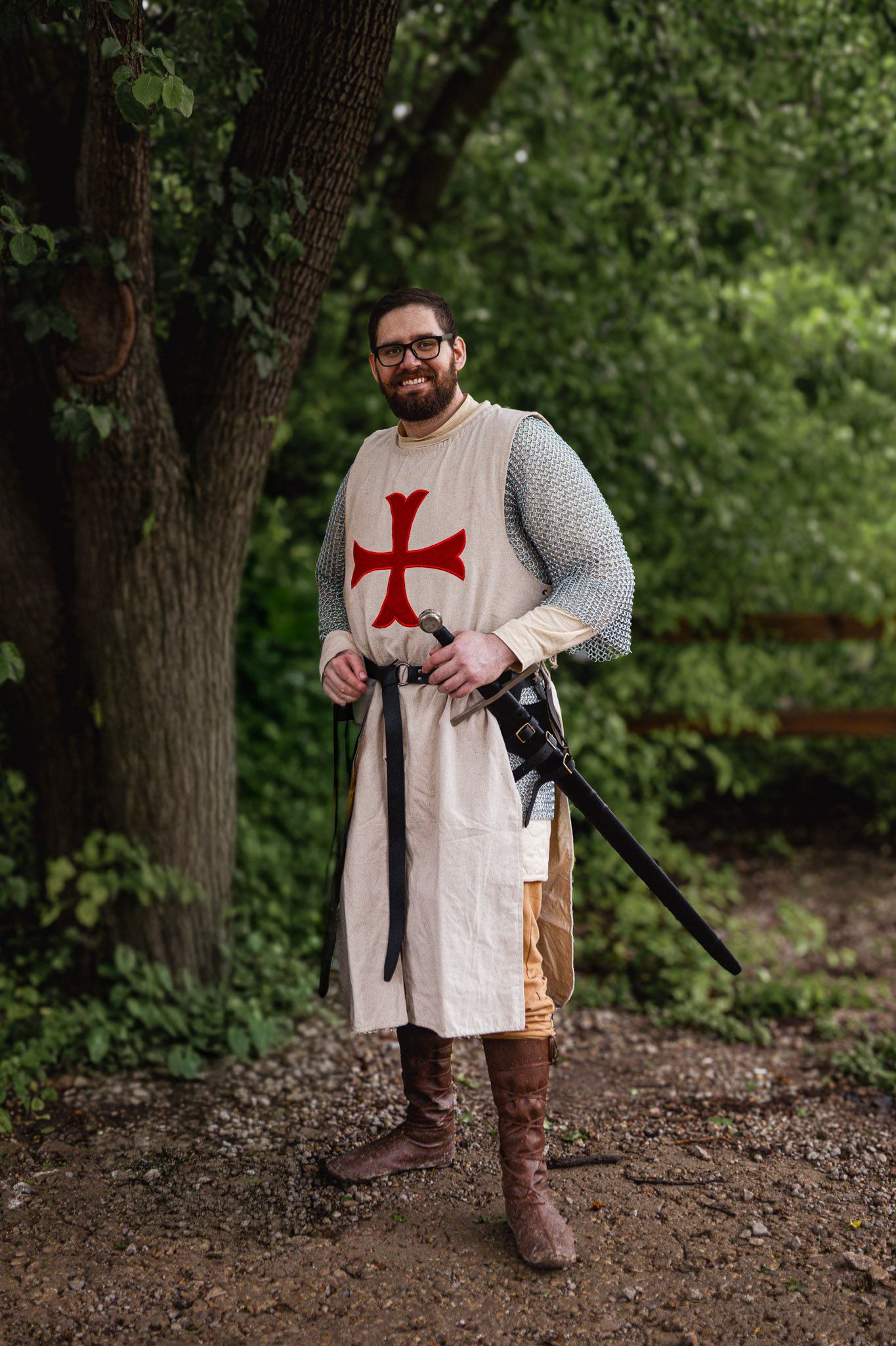 Man in medieval Crusader costume with red cross tabard, chainmail, sword, and boots standing in a forest.