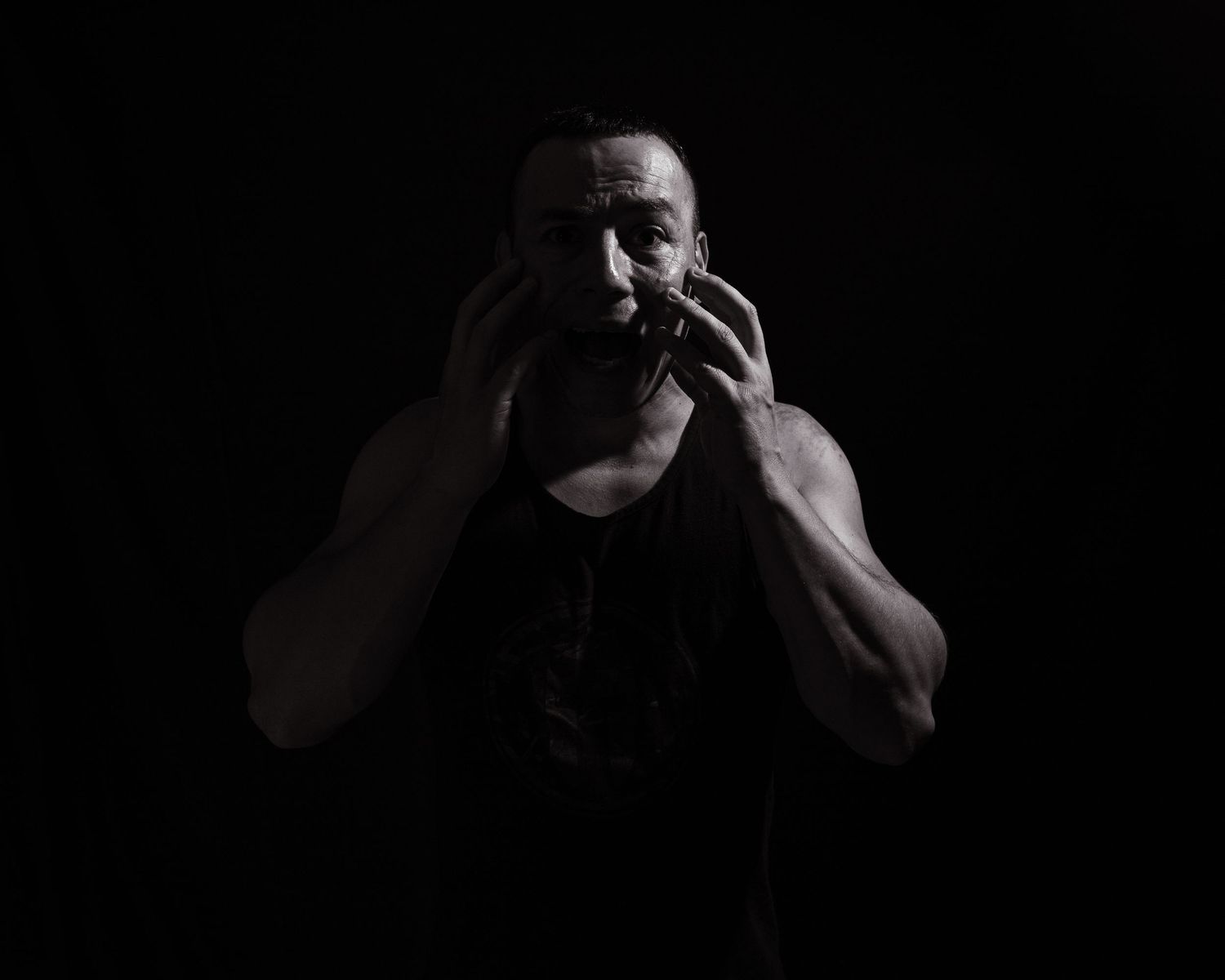 A muscular man in a black tank top holds his face dramatically against a dark background.