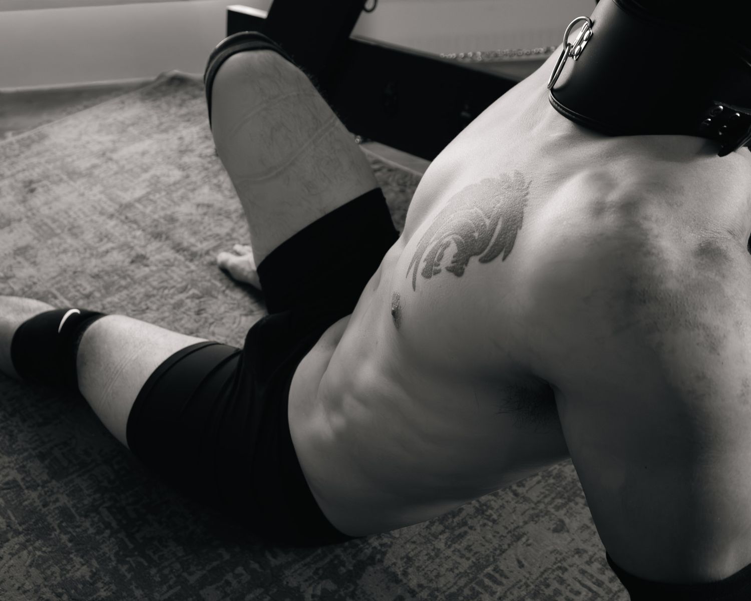 Black and white photo of a shirtless man with tattoo lying on floor wearing black shorts and a collar.