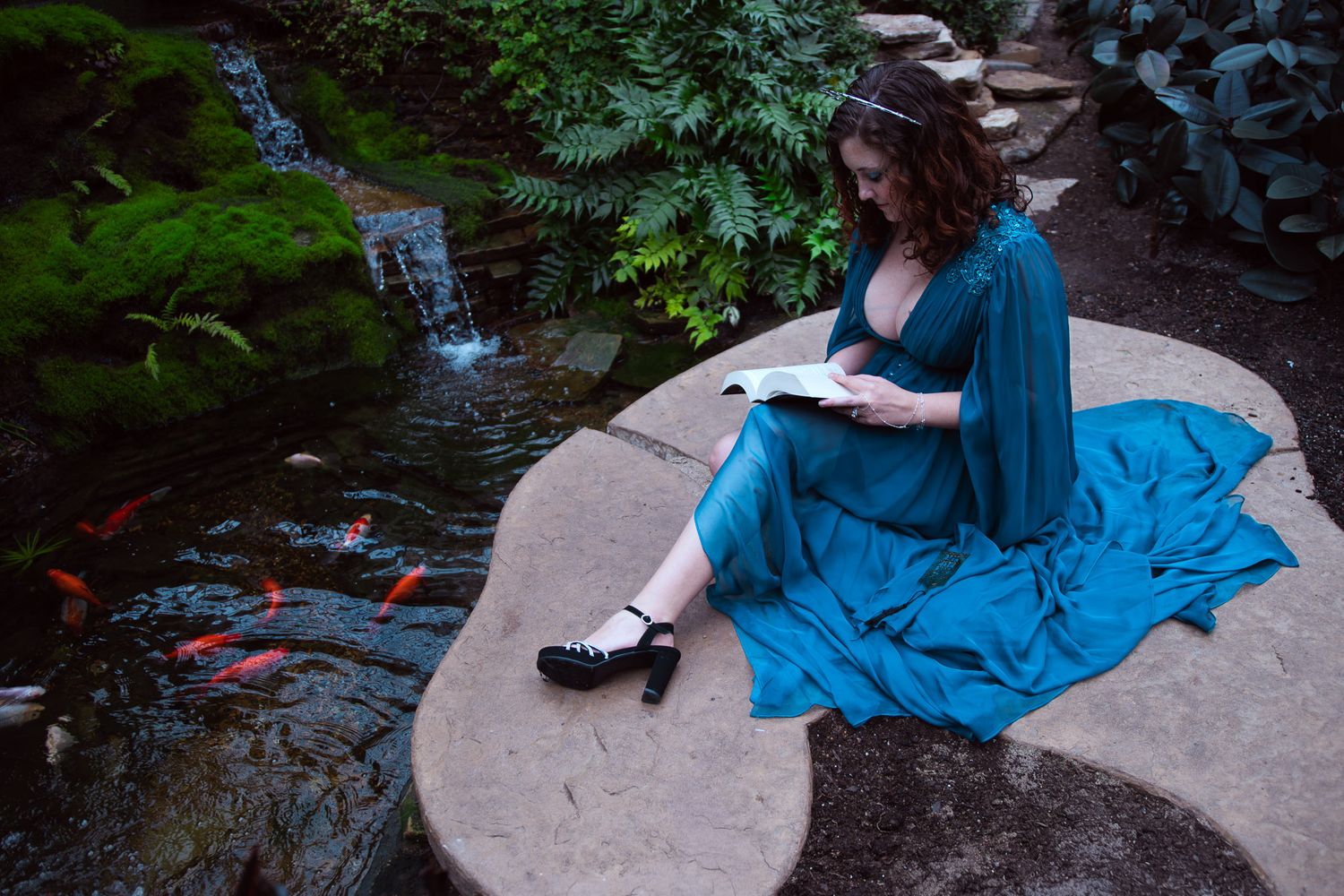 Woman in teal dress reads book beside koi pond with waterfall in lush garden setting.