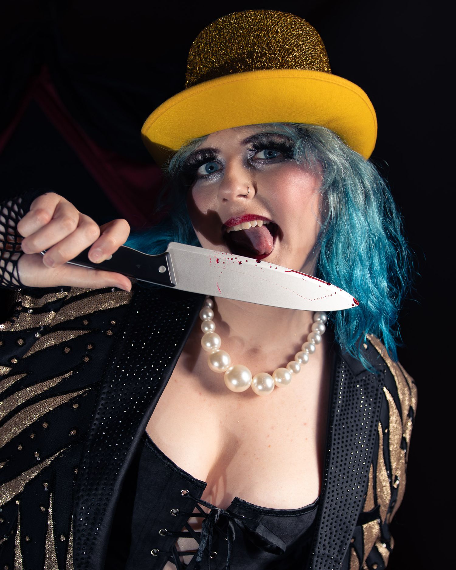 Woman with blue hair and yellow hat licking a knife, wearing pearls and sparkly jacket in dramatic stage performer look.
