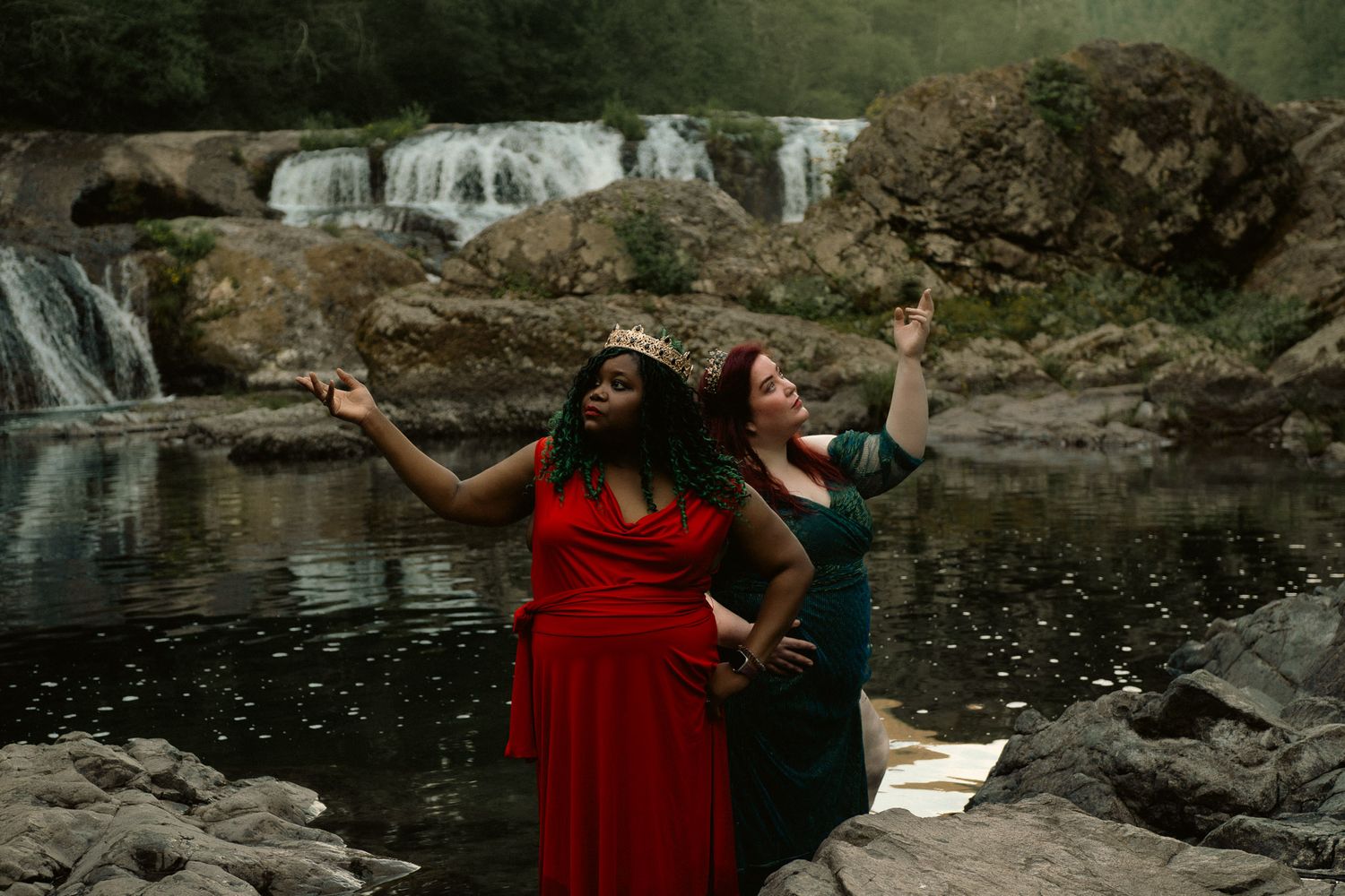 Two women in elegant dresses pose dramatically by a waterfall on rocky terrain in a mystical outdoor setting.