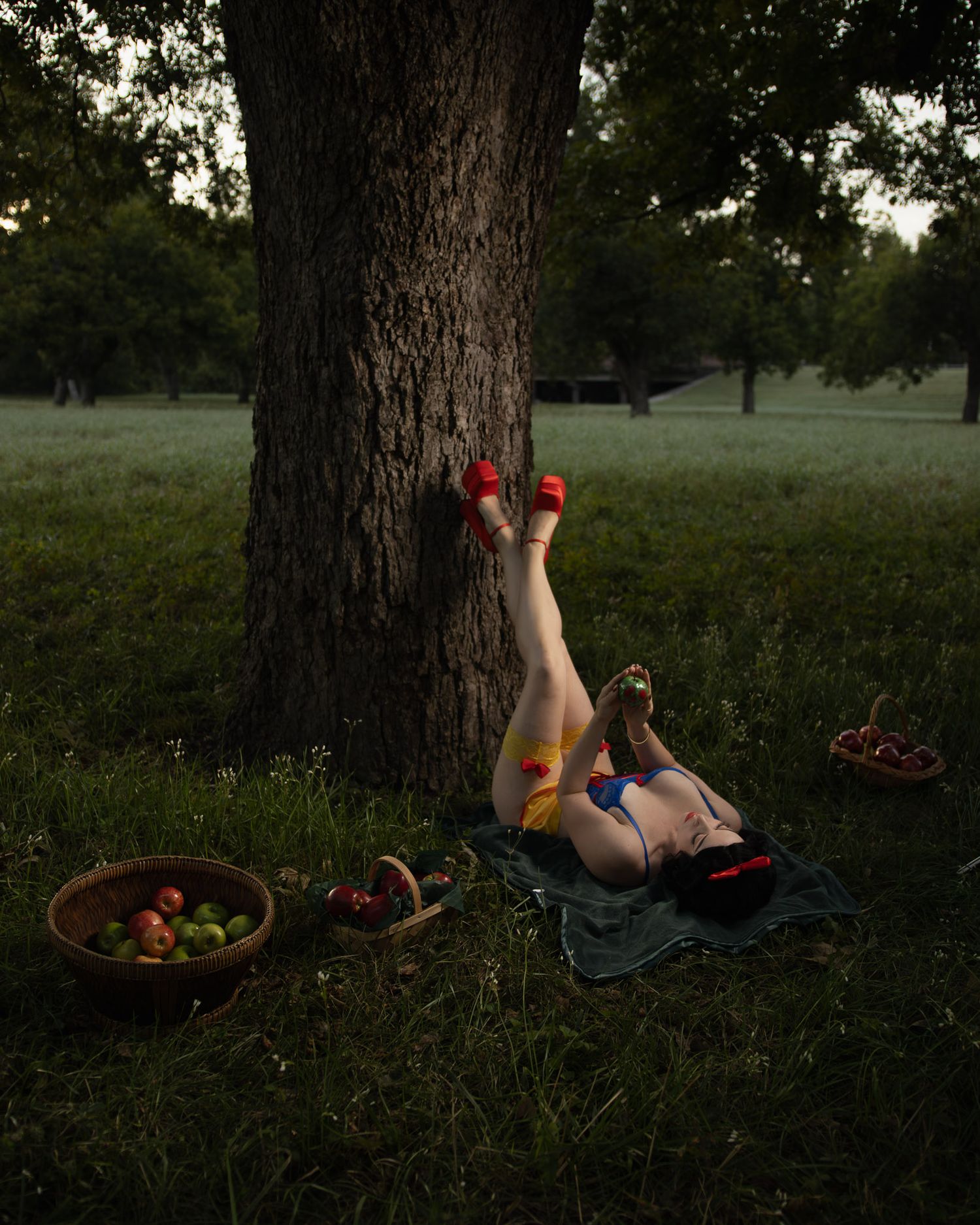Woman in red heels lying on a picnic blanket with her legs propped against a large tree, baskets of apples nearby in a dusky park.