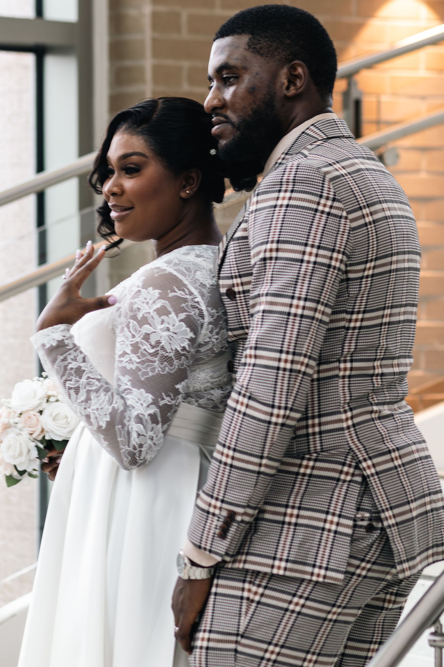 Bride in lace wedding gown holds bouquet beside groom in plaid suit on elegant staircase.