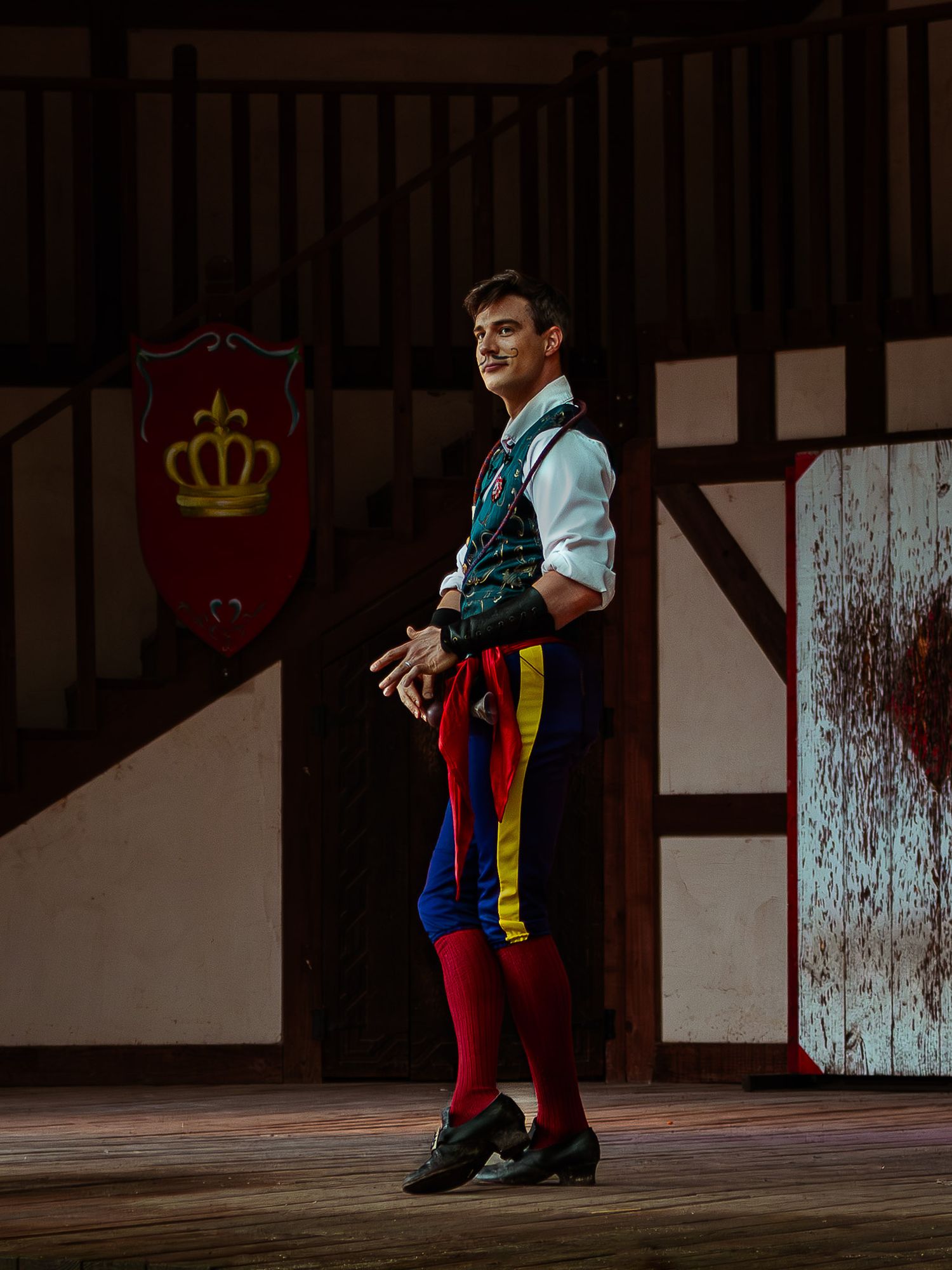 Male performer in colorful medieval costume on stage with crown shield backdrop.