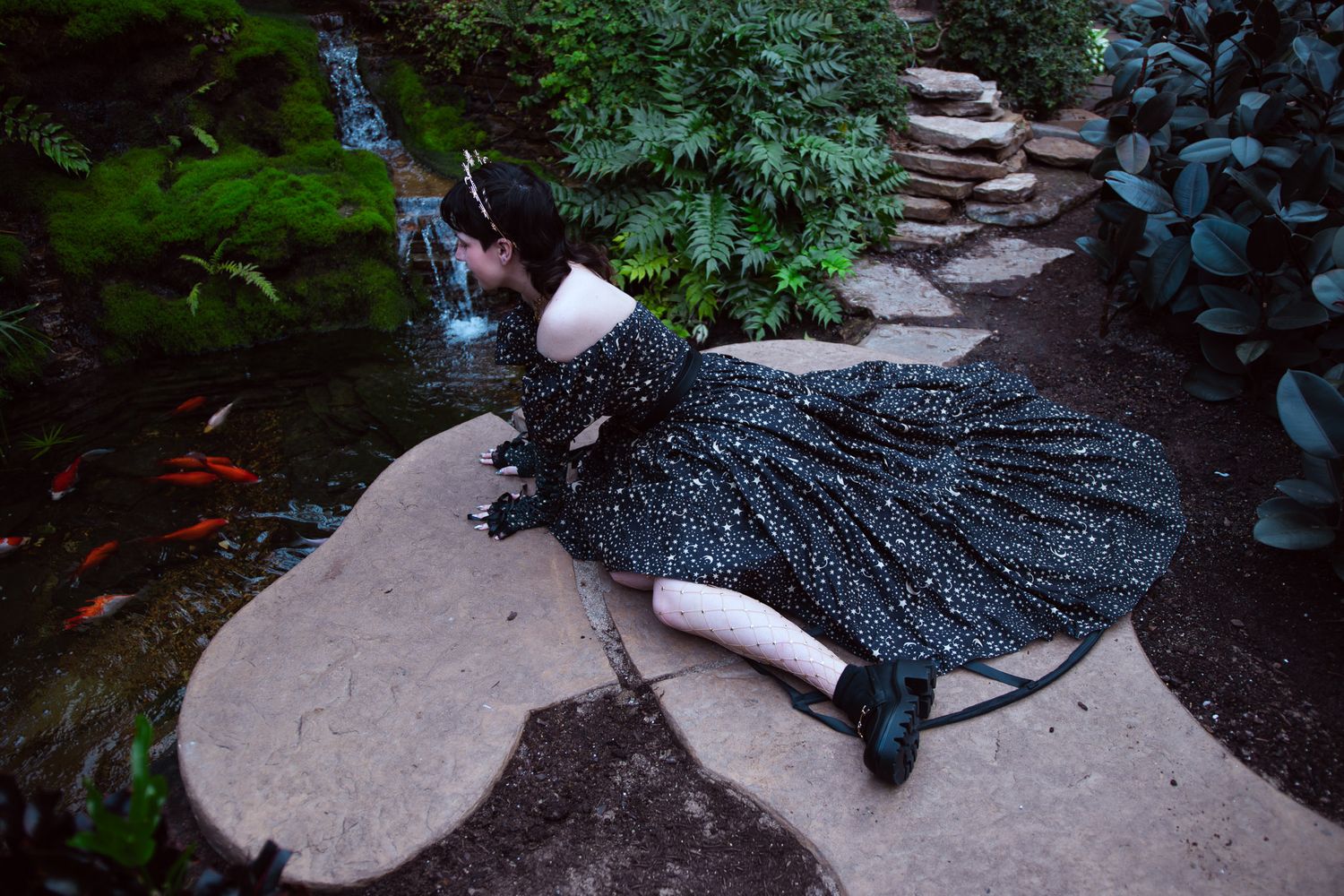 Girl in black starry gown sits by a koi pond in a lush garden with mossy rocks and ferns.