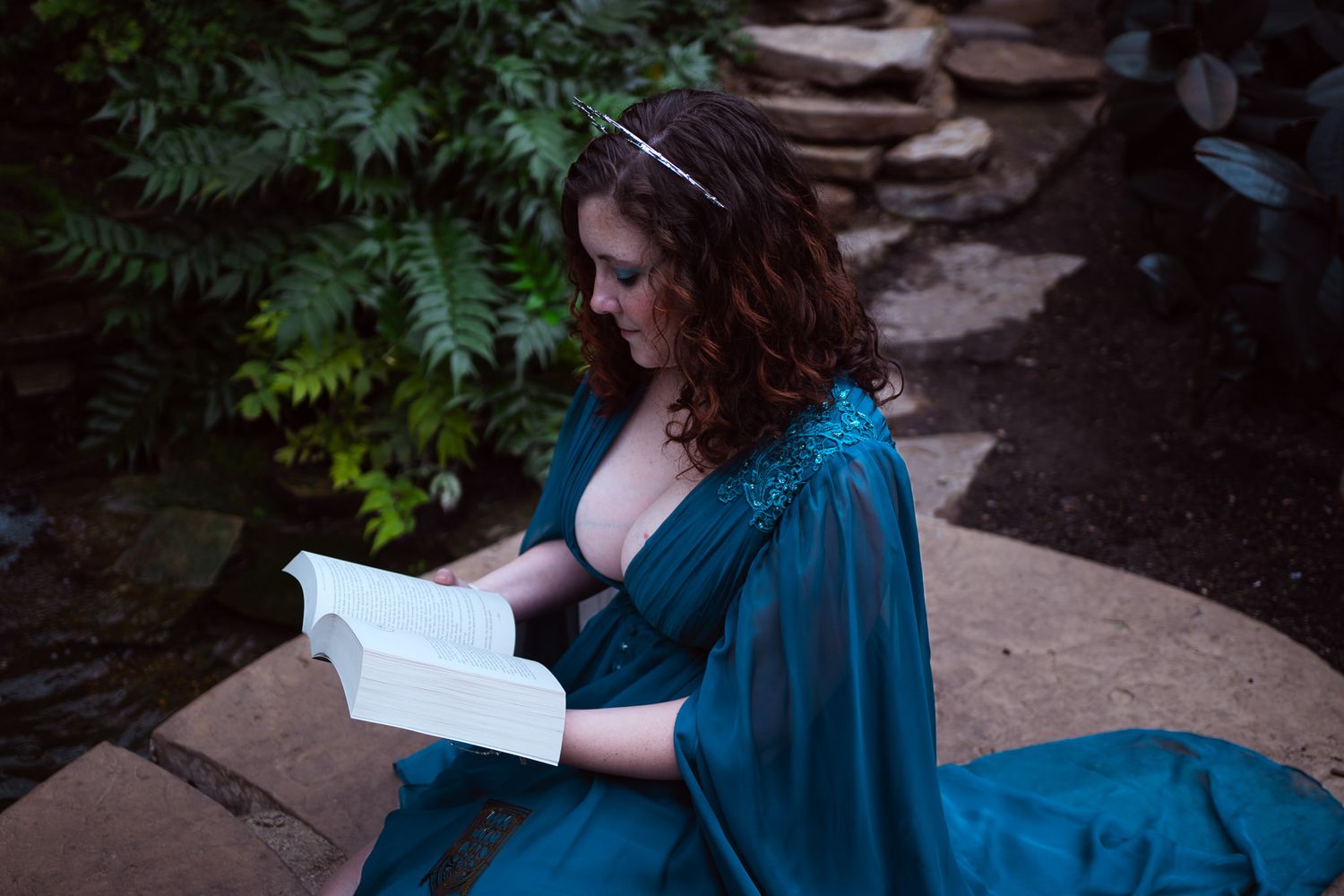 Woman in teal gown reading a book outdoors among lush greenery and stone steps.