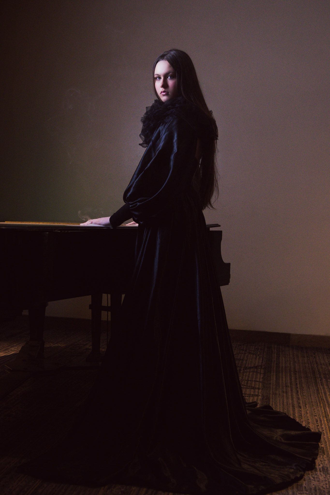 Woman in long black velvet gown standing by a grand piano in a dimly lit room, looking over her shoulder.
