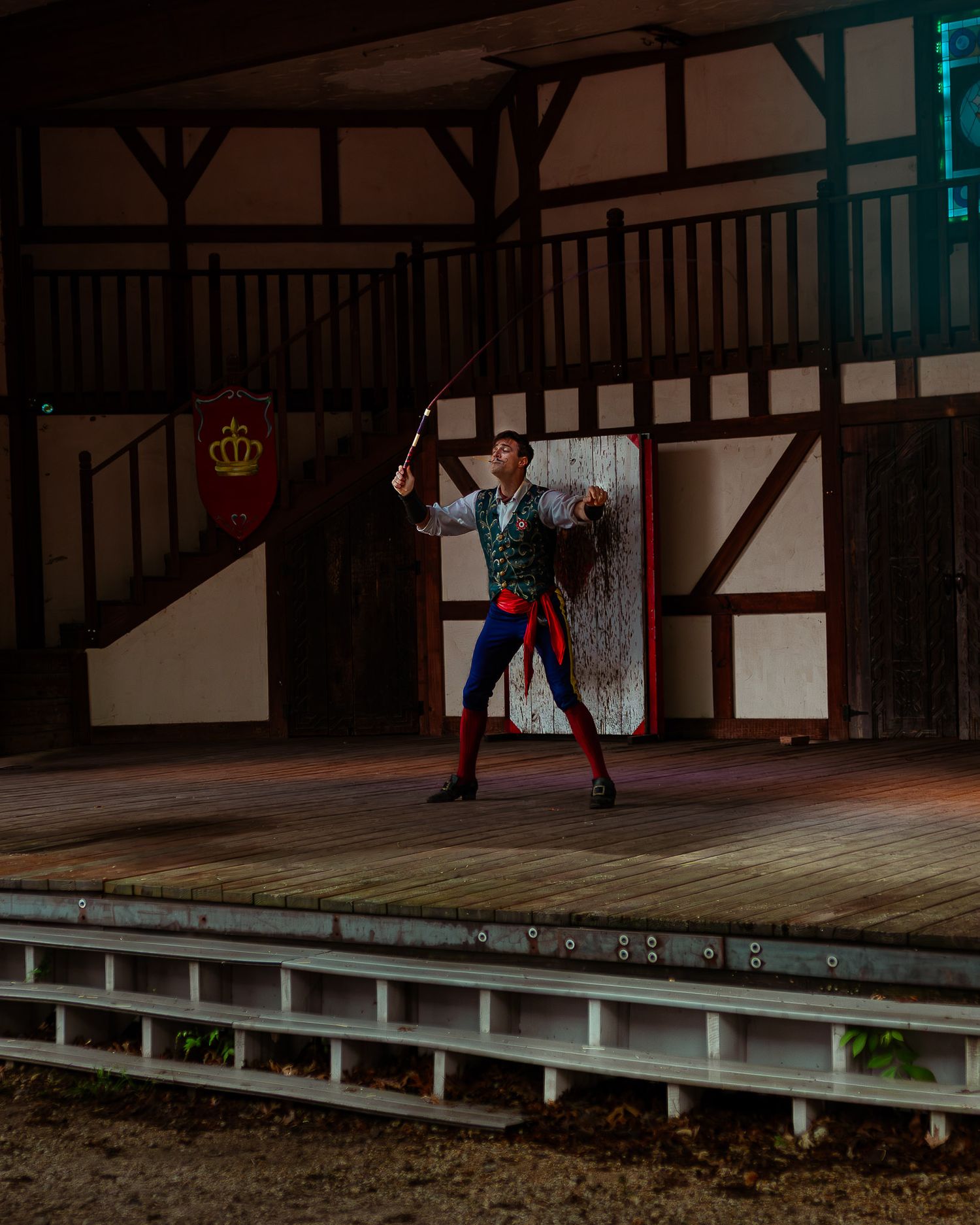 A performer in medieval costume stands on an outdoor stage holding a whip at a Renaissance fair.