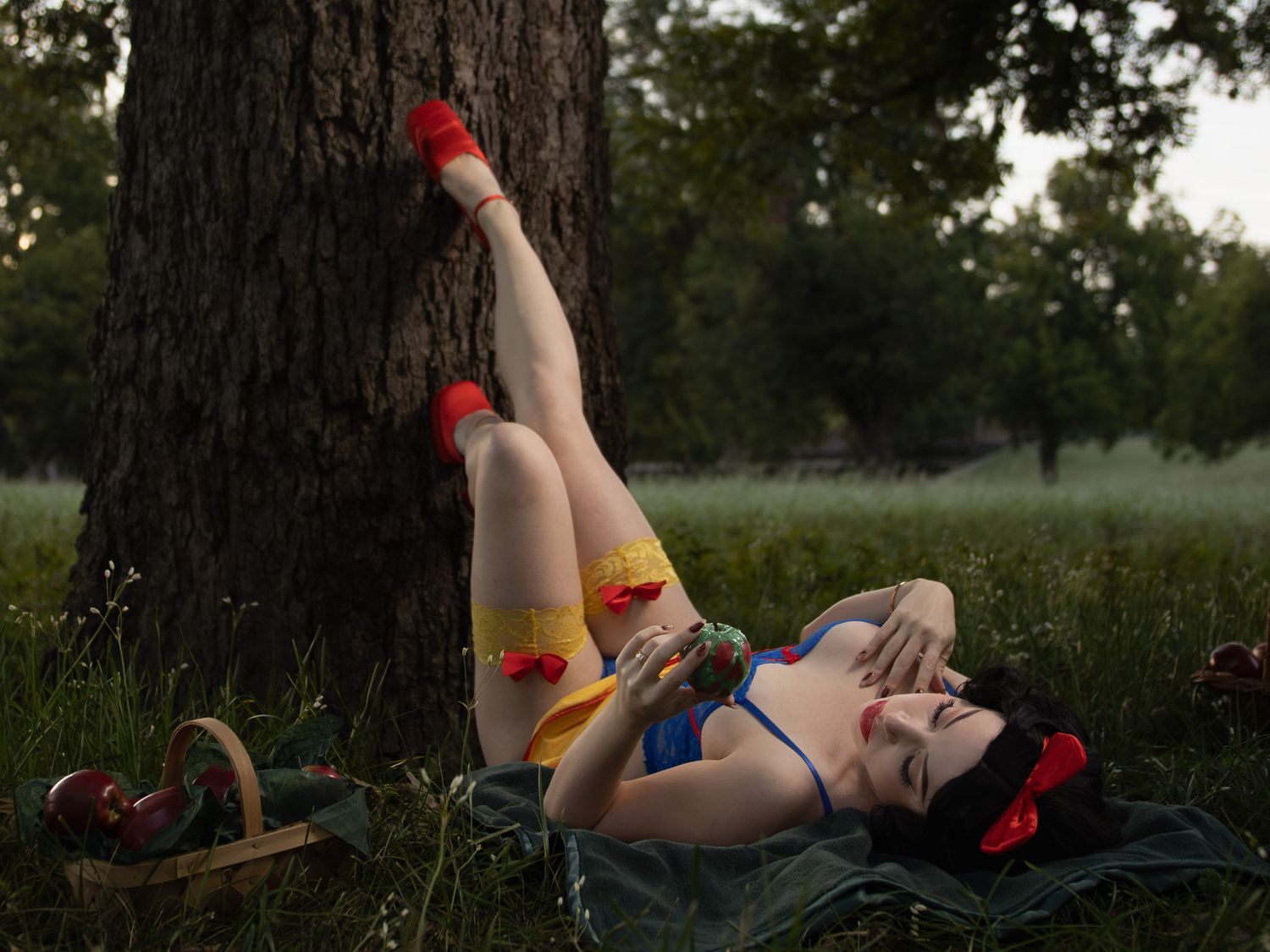 Woman in a Snow White-inspired costume lies on a blanket in a grassy park, legs propped against a tree trunk, holding a red apple.