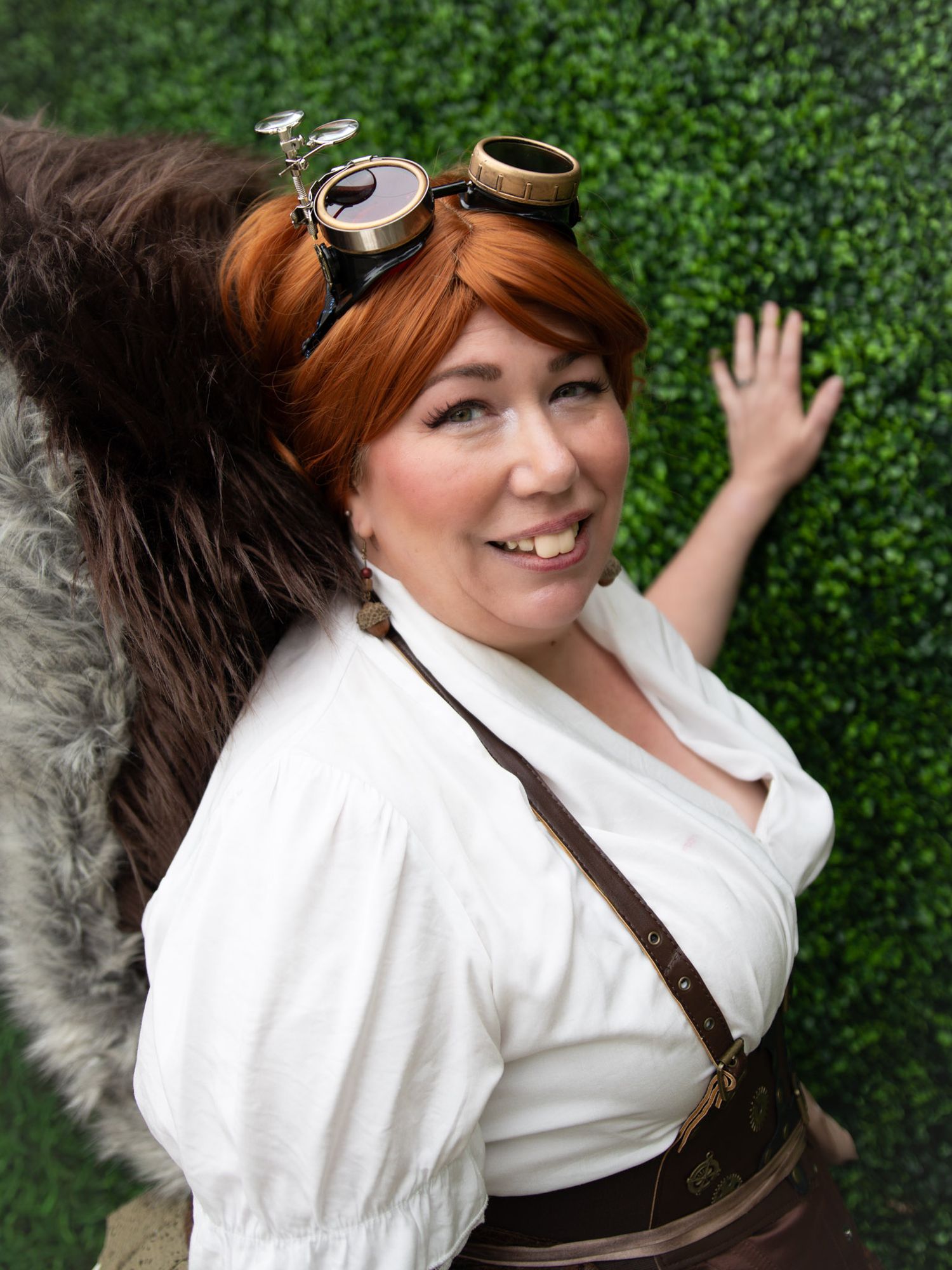 Woman in steampunk cosplay with goggles, white blouse, brown corset, and fur accessory poses before a green hedge wall.
