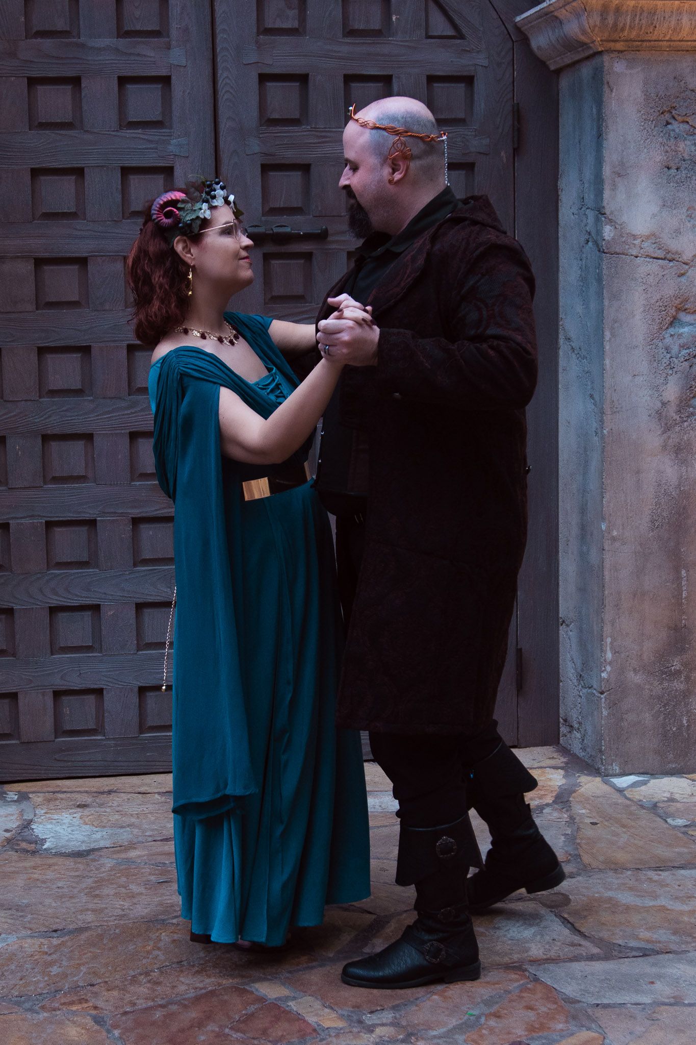 Couple in medieval costumes dancing, woman in teal gown with floral crown, man in black attire, stone courtyard.