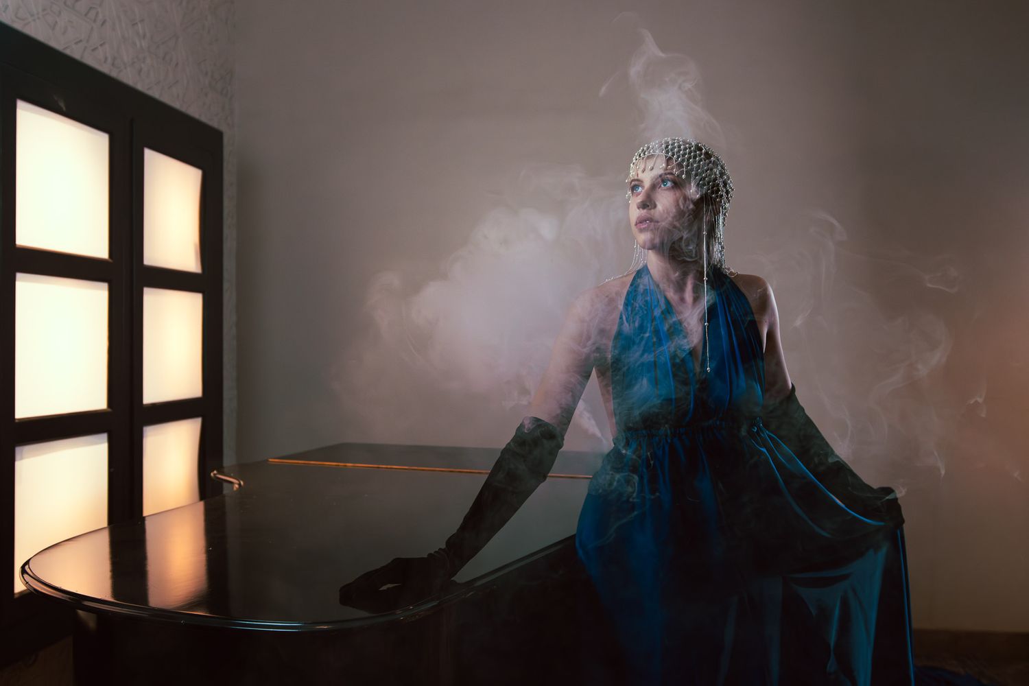 Woman in blue gown and pearl headpiece poses dramatically with smoke swirling around her in a dimly lit room.