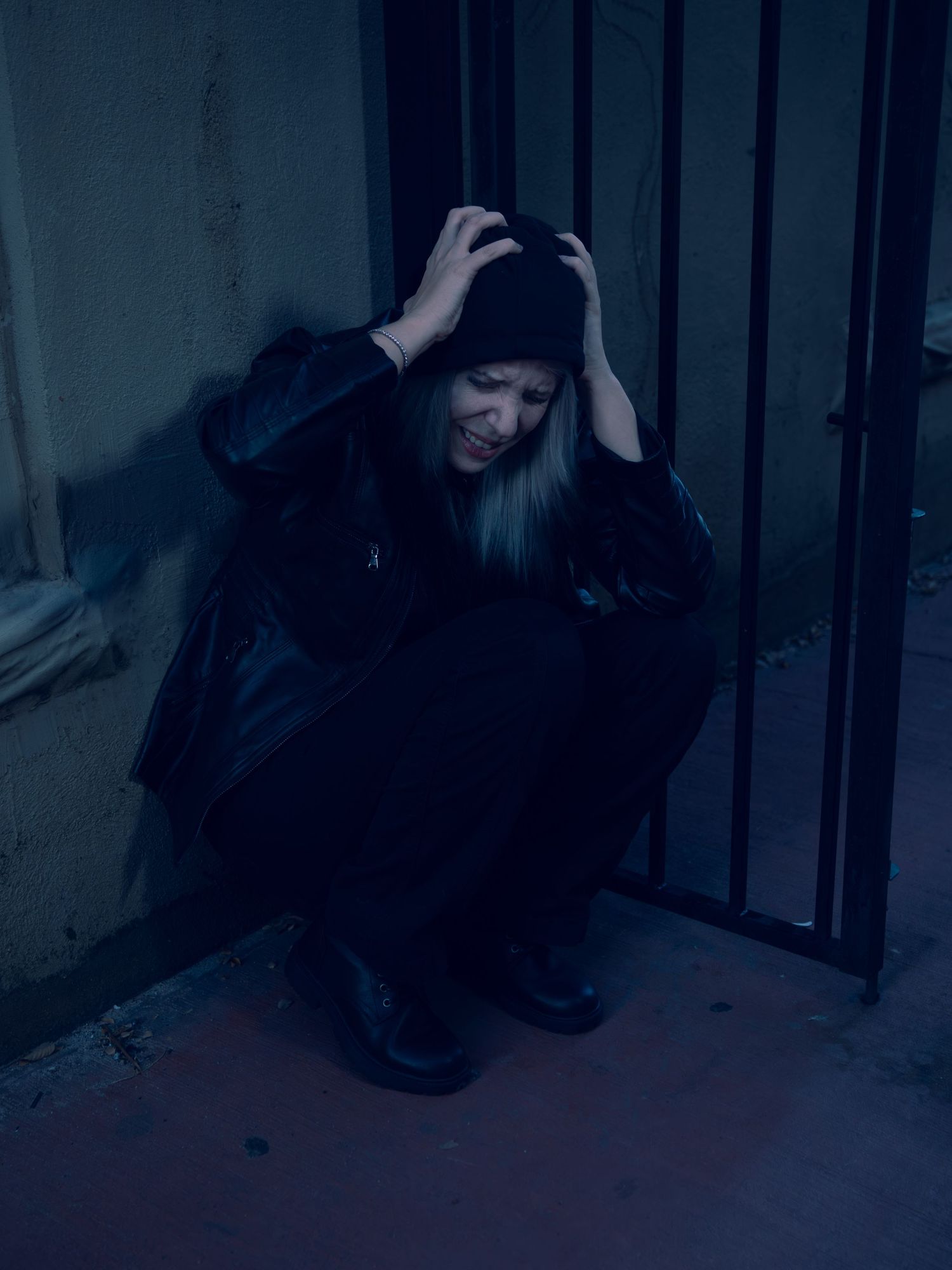 A distressed woman in black crouches against a dark wall by metal bars, hands clutching her head in despair.