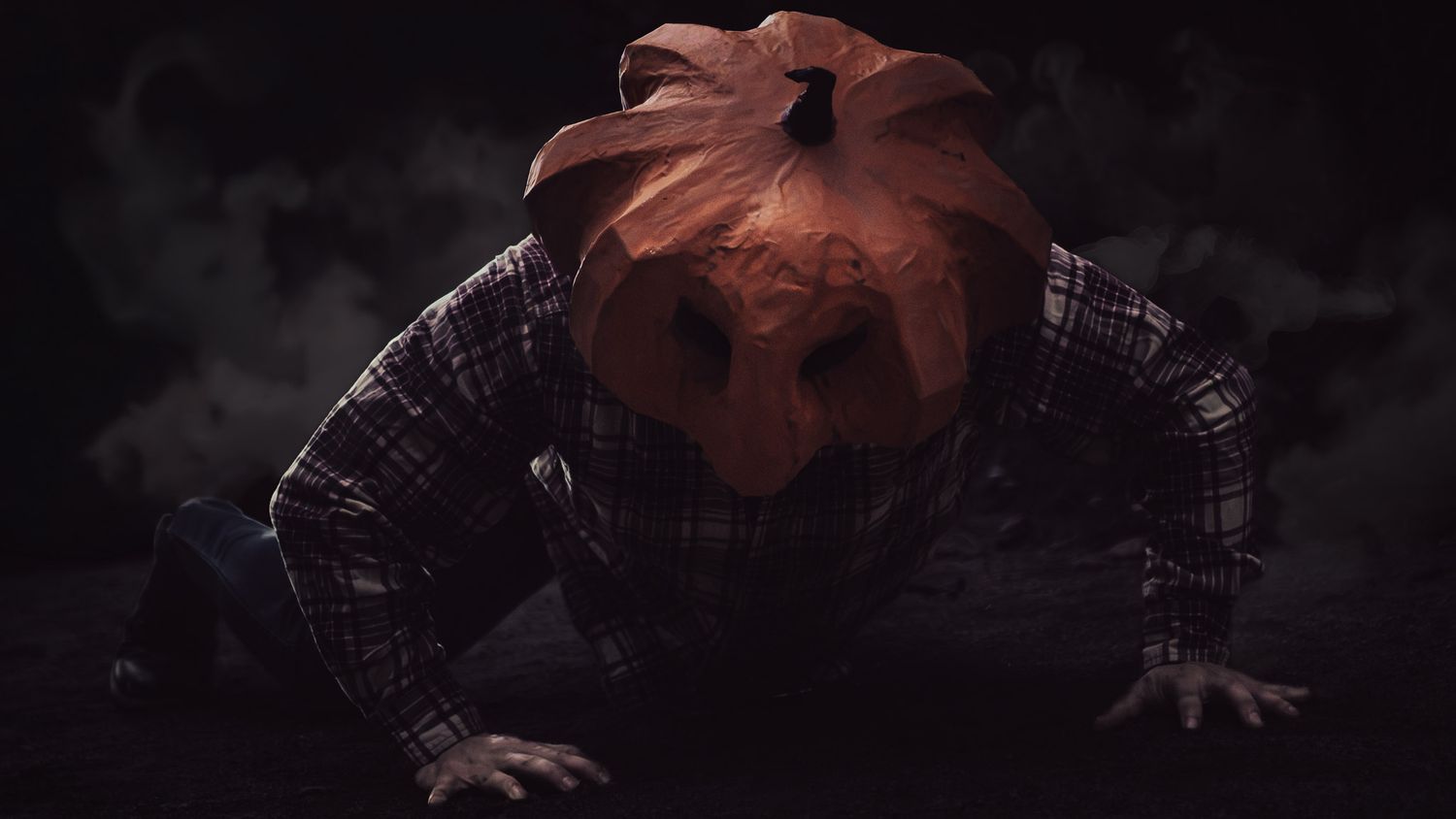 Creepy Halloween figure with jack-o-lantern pumpkin head crawling in dark smoky atmosphere.