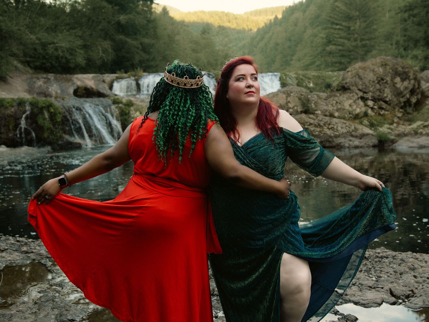 Two plus-size women in elegant red and green gowns pose by a waterfall in a lush forest setting.