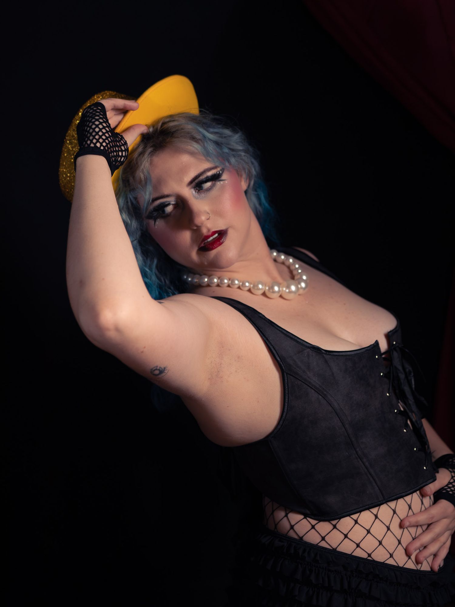 Woman with blue hair in black corset and pearl necklace poses dramatically holding yellow hat in dark studio.