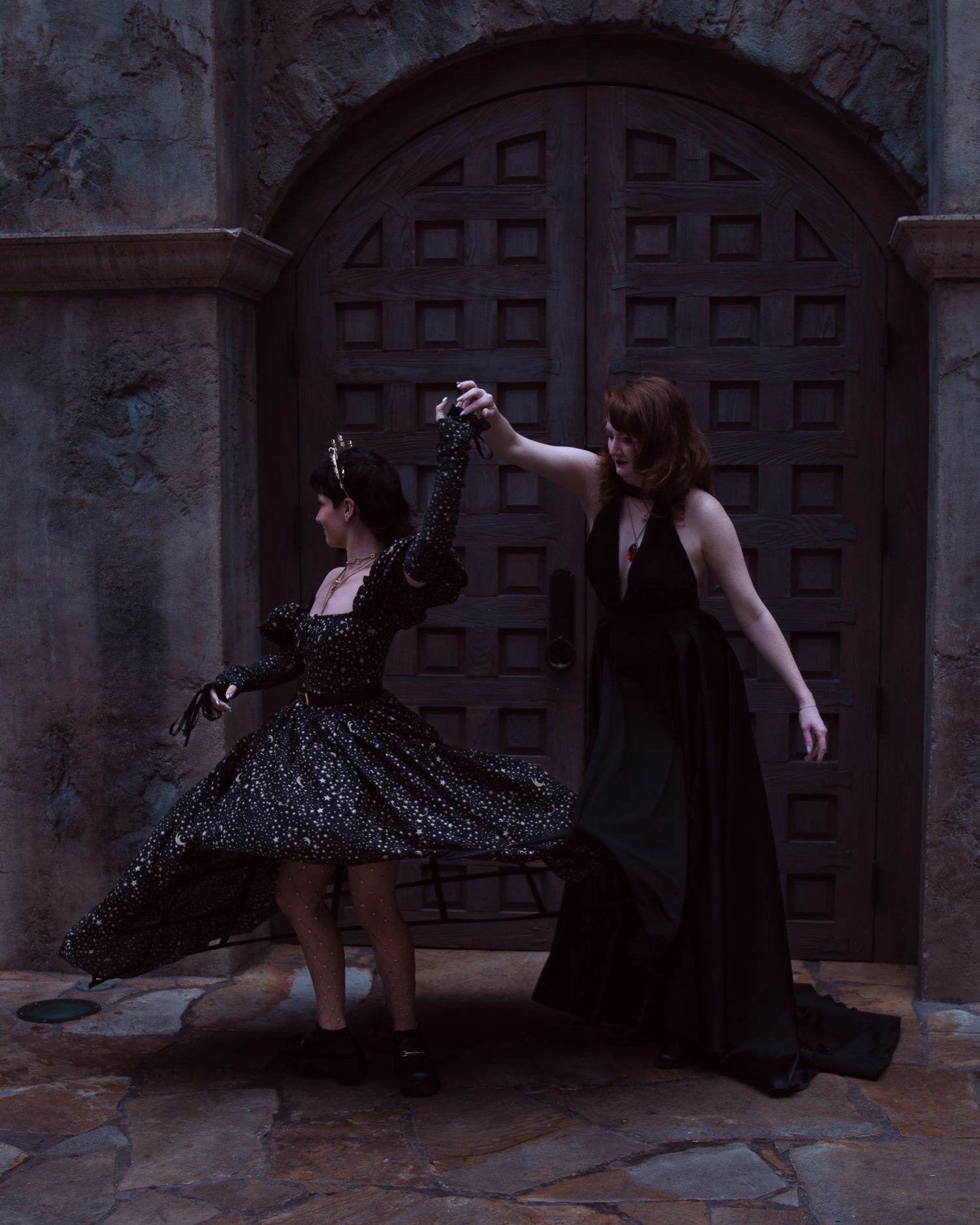 Two women in elegant black gowns dancing dramatically before ornate wooden doors in a gothic setting.
