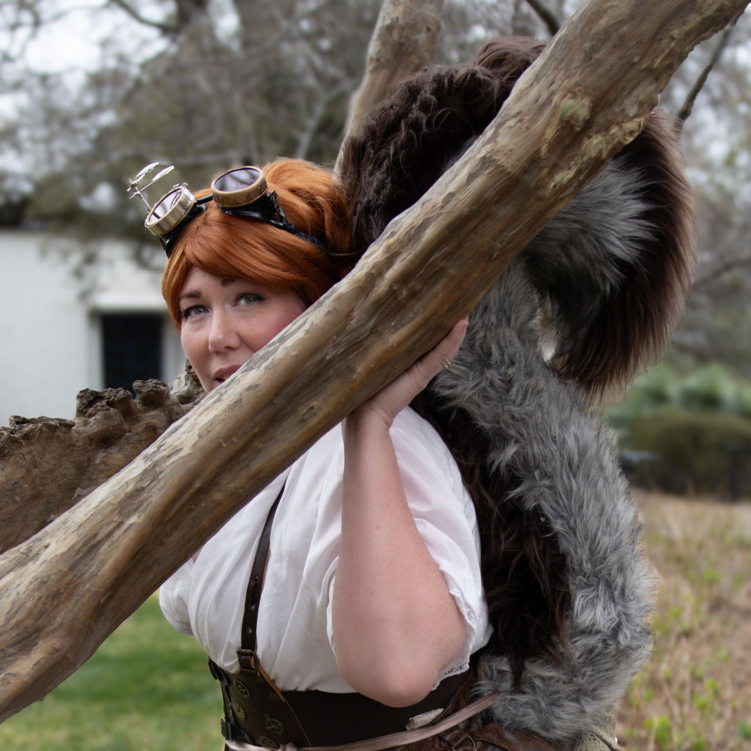 Woman in steampunk costume with goggles and fur accessory posing behind a large tree branch outdoors.