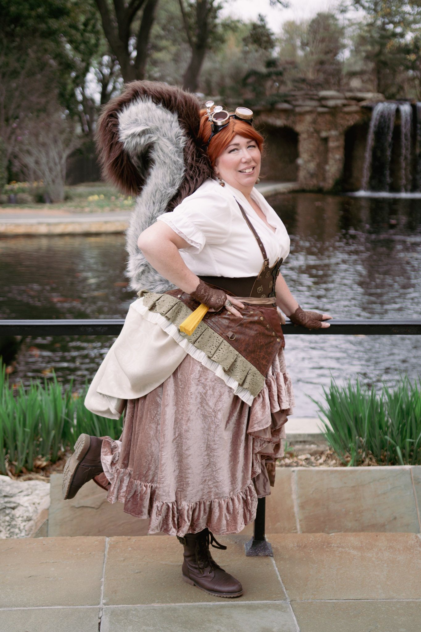 Woman in steampunk costume with squirrel tail, goggles, ruffled skirt, and boots poses outdoors near a waterfall.