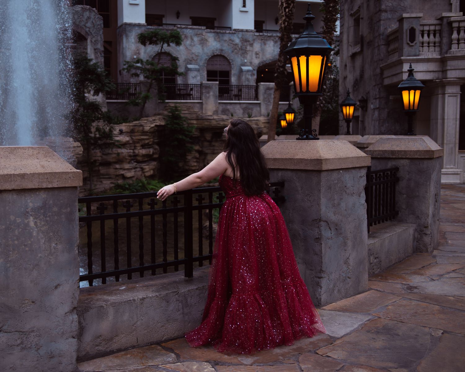 A girl in a sparkling red ballgown stands near a stone railing with glowing lanterns in an elegant courtyard.