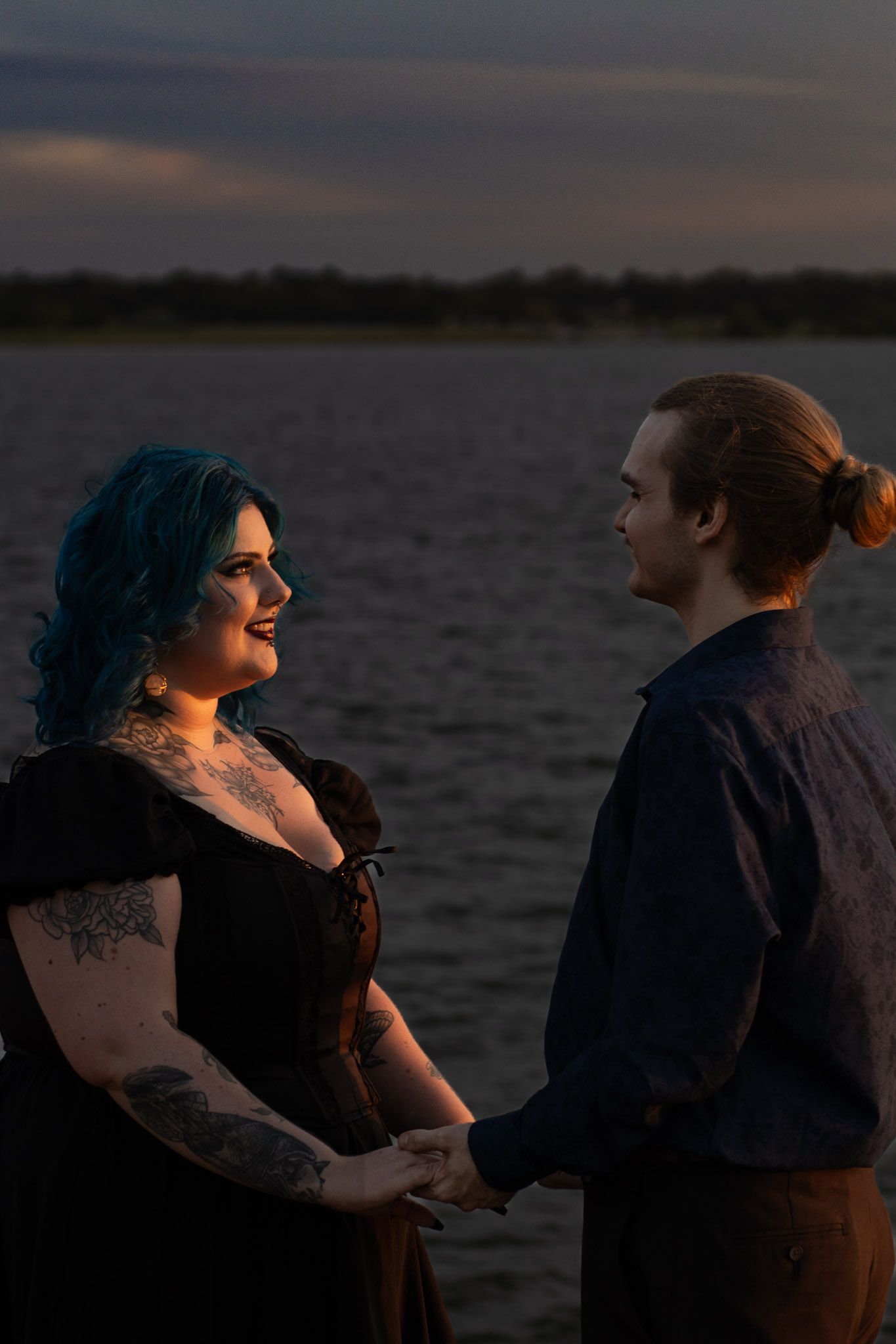 Couple holding hands at dusk by a lake, woman with blue hair and tattoos in black dress, man with bun hairstyle.