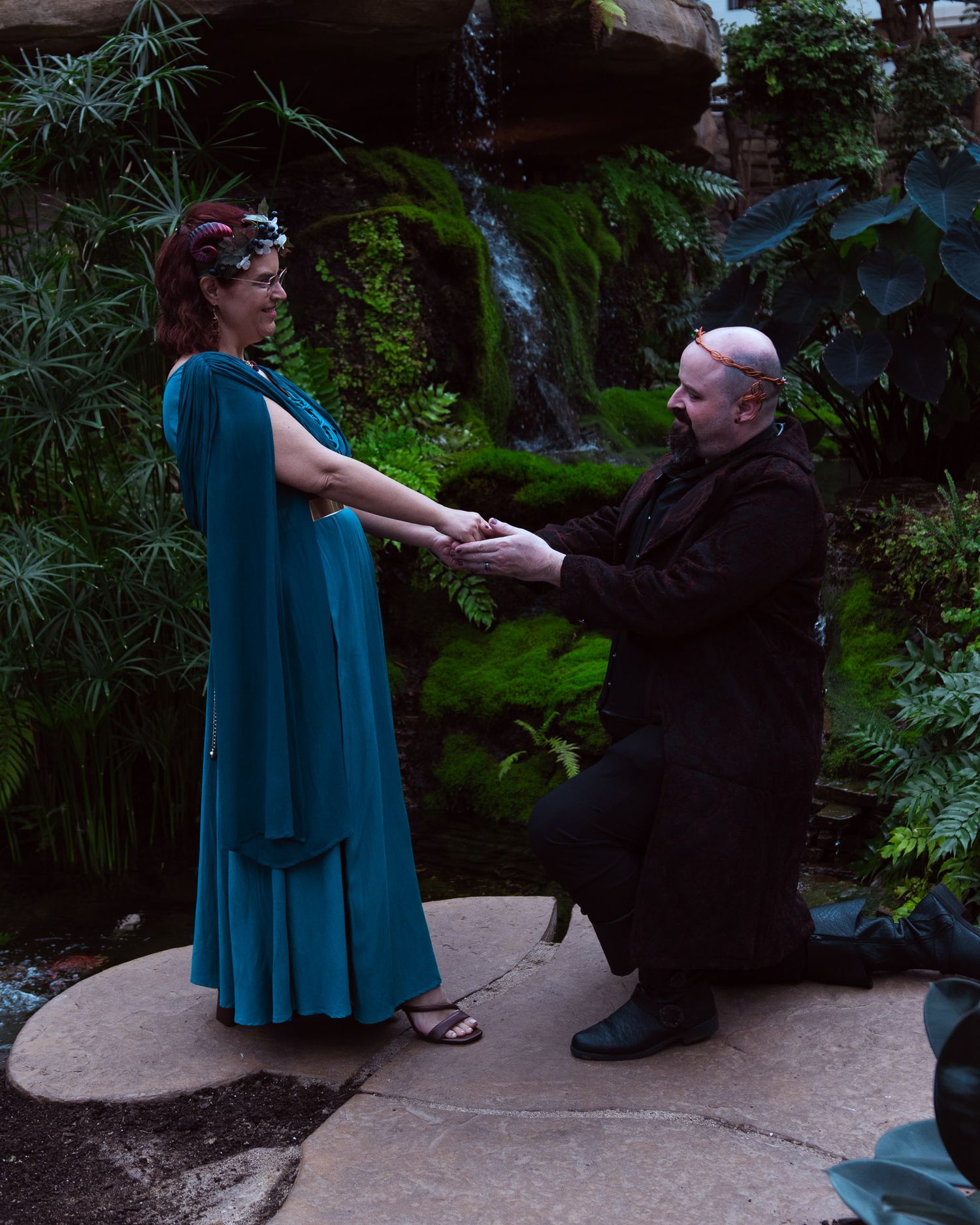 Man kneeling before woman in teal dress during romantic garden proposal near waterfall.