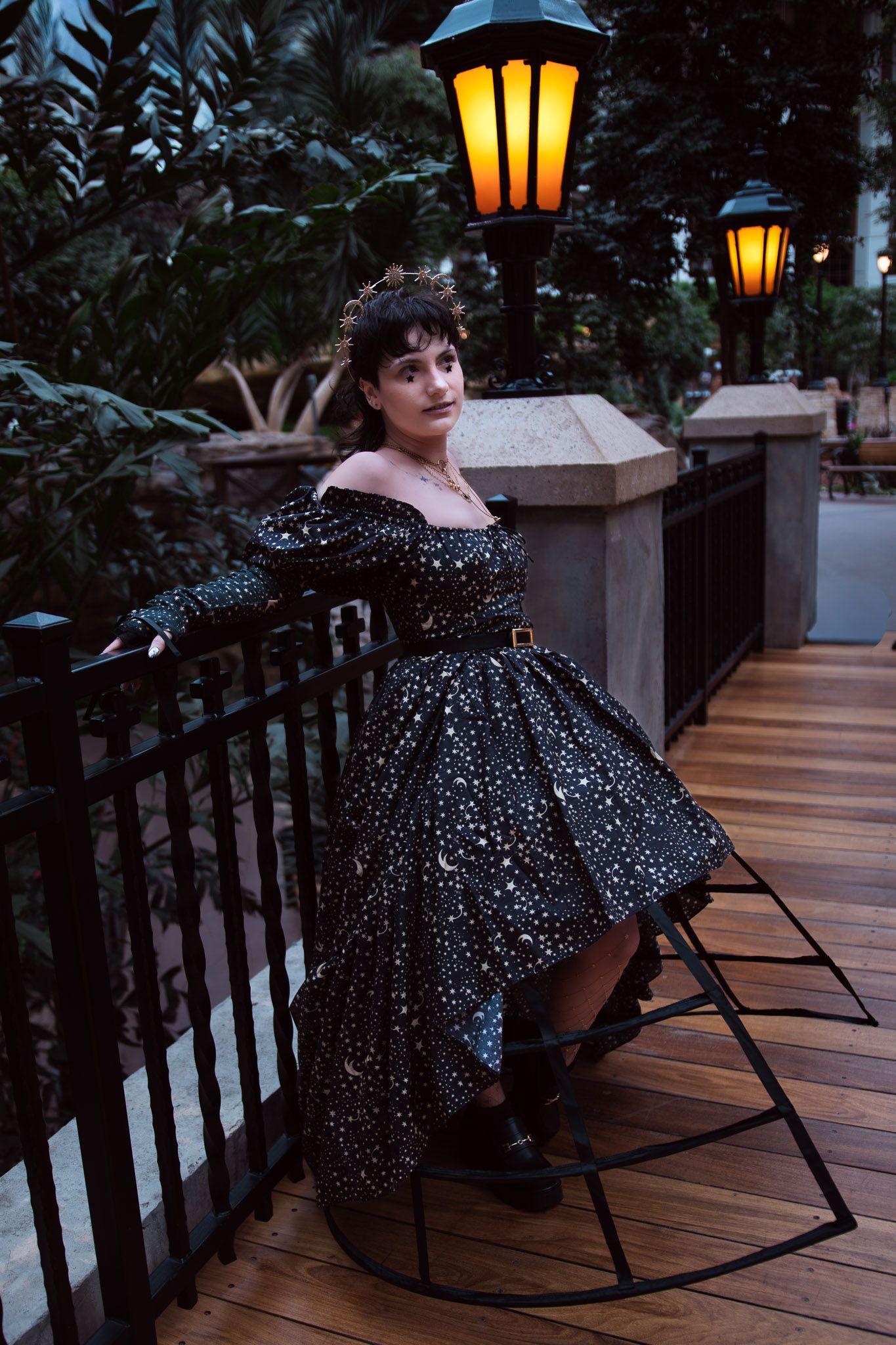 Woman in black celestial off-shoulder gown poses on wooden bridge with glowing lanterns and lush greenery.