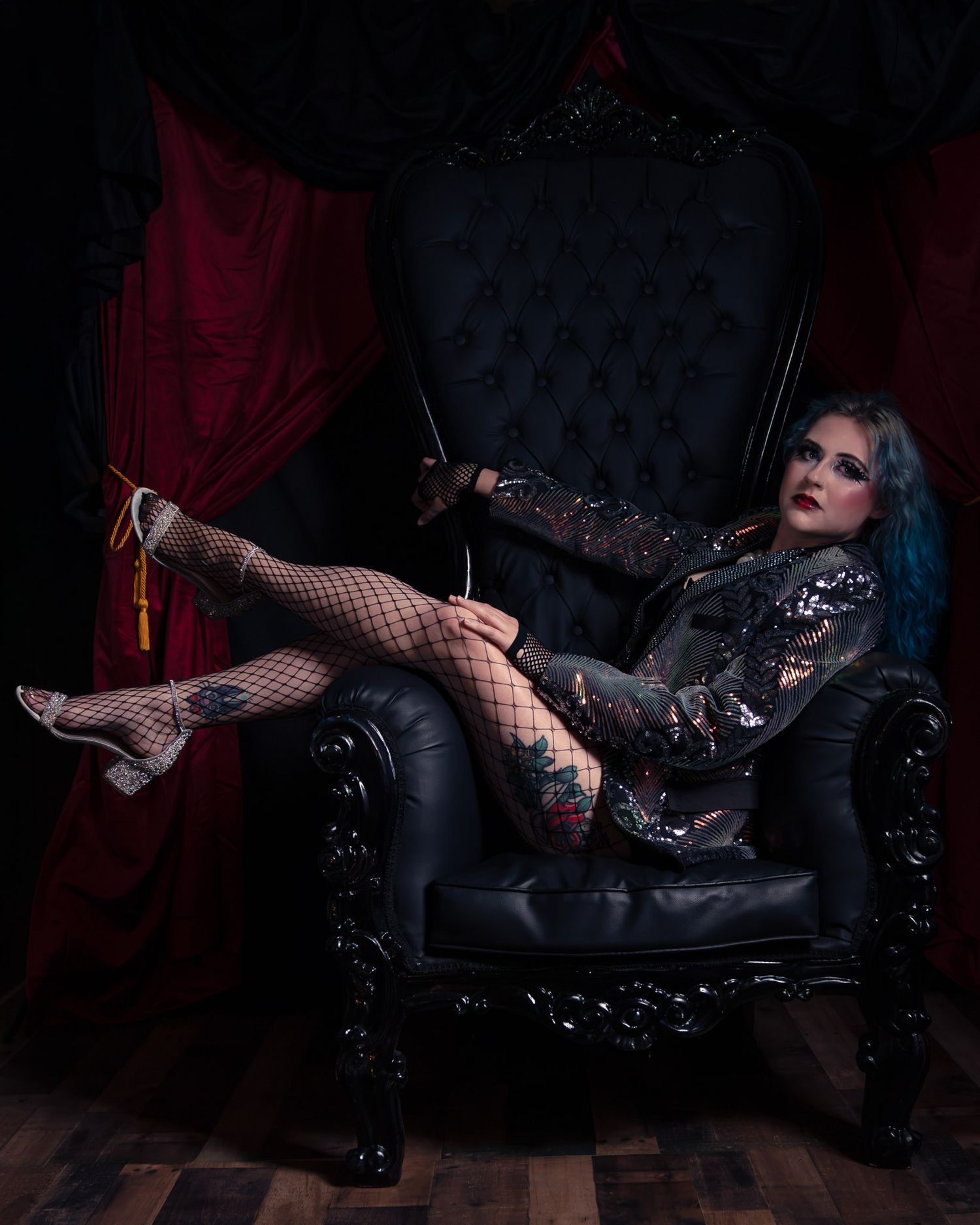 Gothic woman with blue hair in fishnet stockings poses on black throne chair with red velvet curtains.