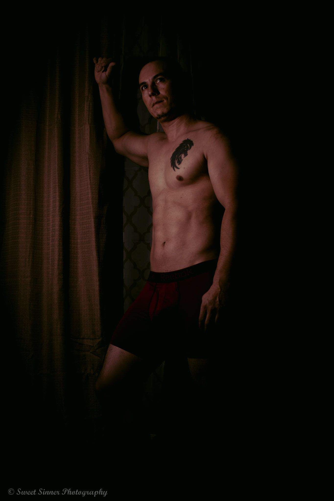 A moody male boudoir portrait of a tattooed man in dark red underwear, posing against a dark, draped curtain background.