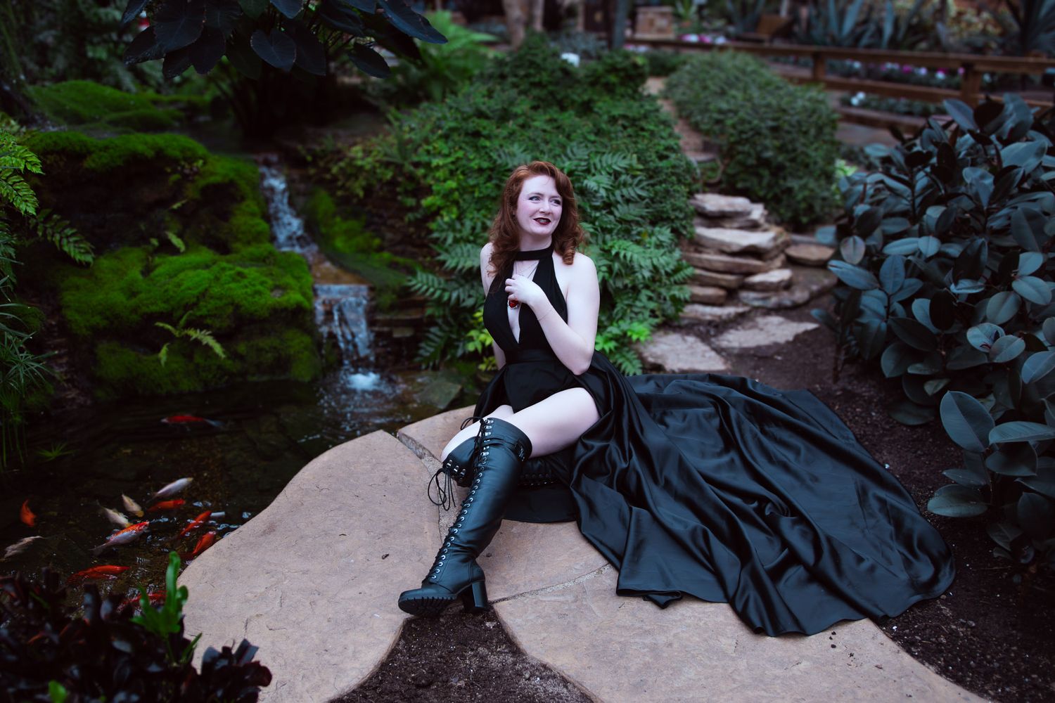 A redhead woman in a black gown and boots sits on a stone in a lush garden with a waterfall.