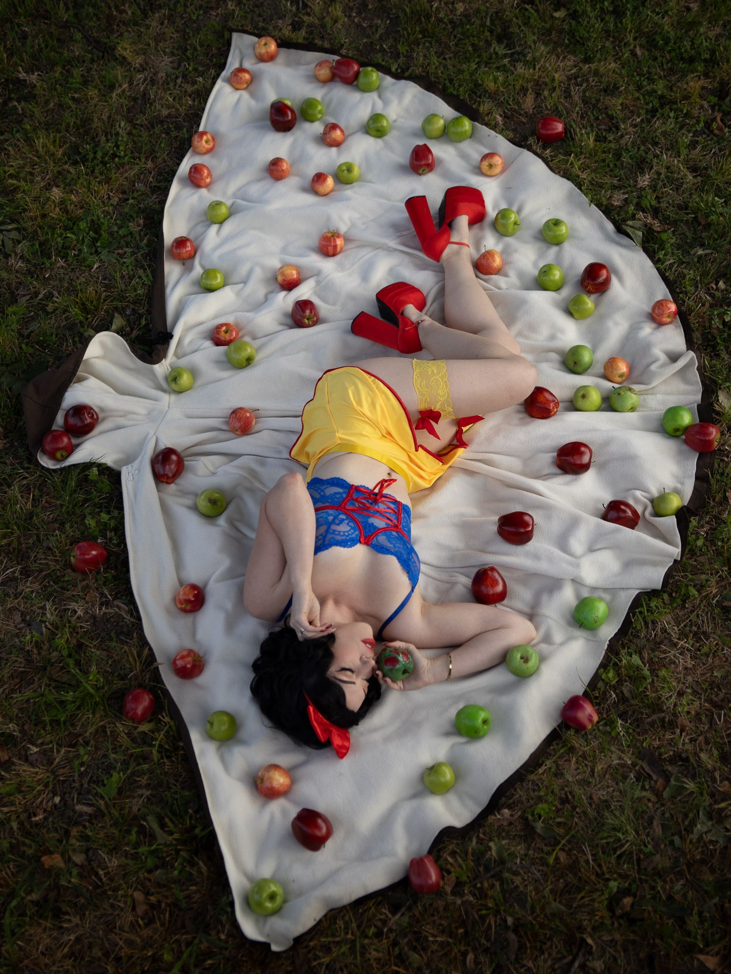 Snow White cosplayer in a yellow skirt and red heels lying on a white cape fanned out over grass and scattered with red and green apples.