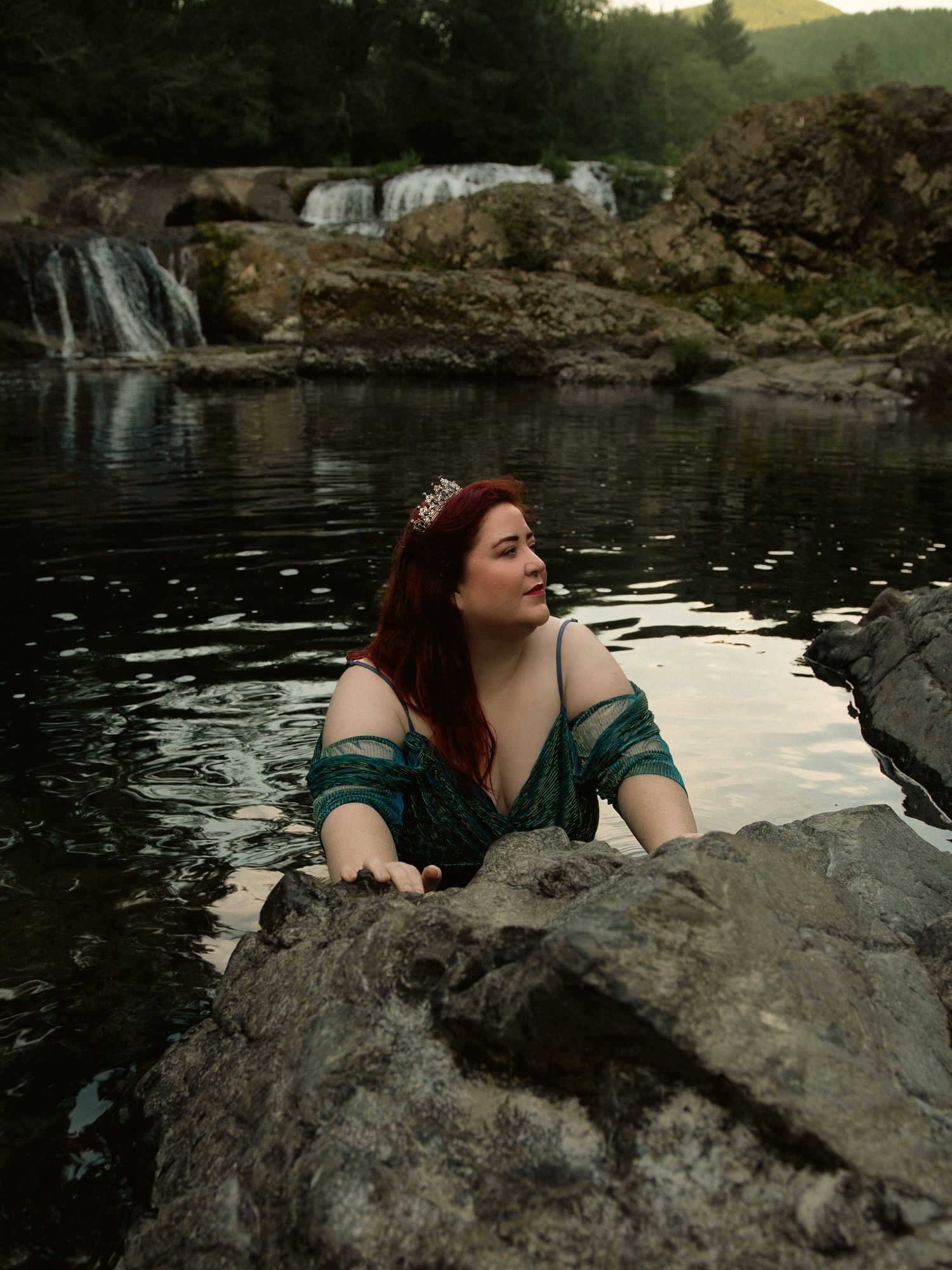 A red-haired woman wearing a green gown and tiara emerges from a rocky river pool with a waterfall backdrop, having her mermaid moment.